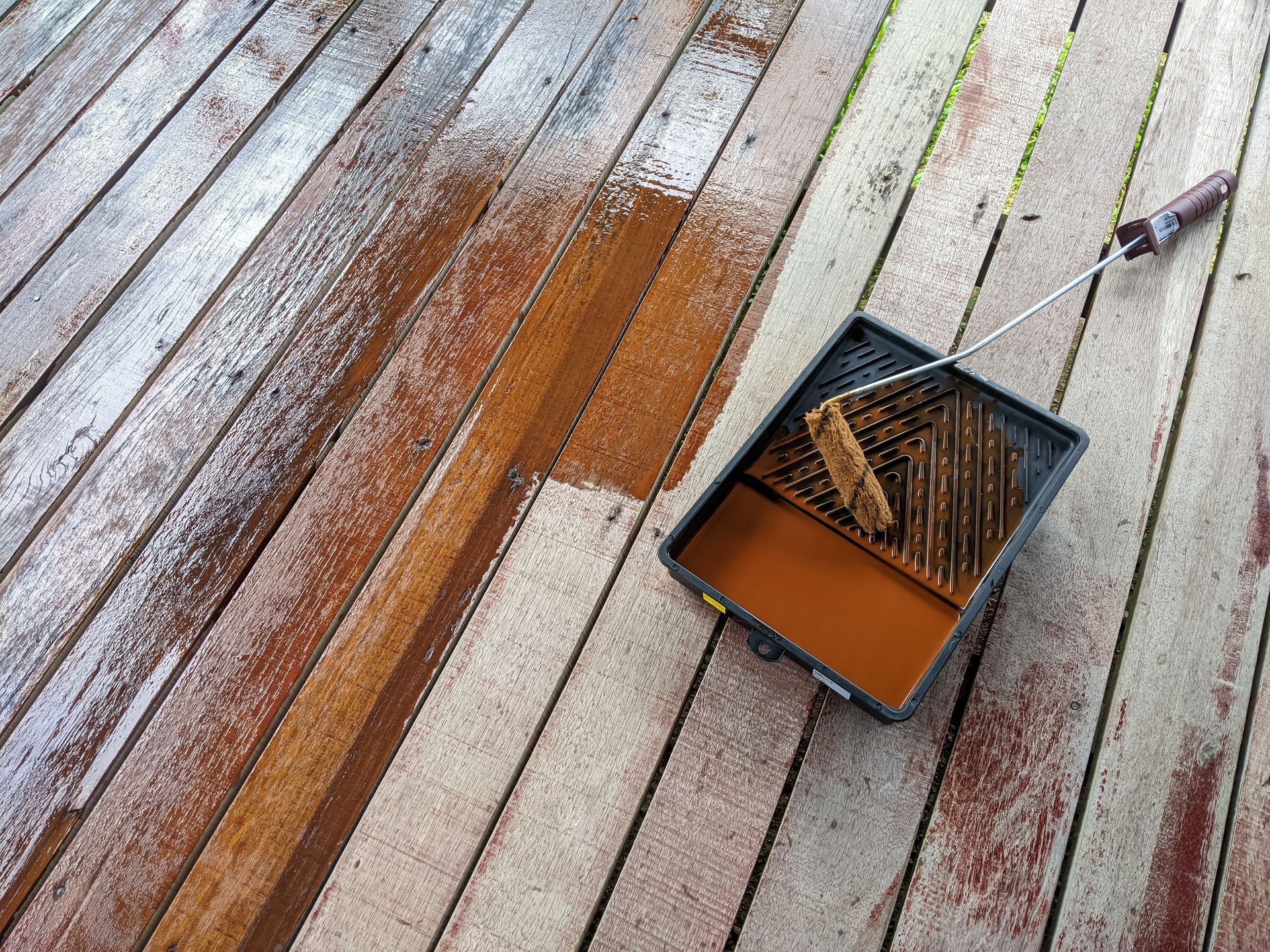 A paint roller and tray filled with brown deck stain sit on a partially stained wooden deck.