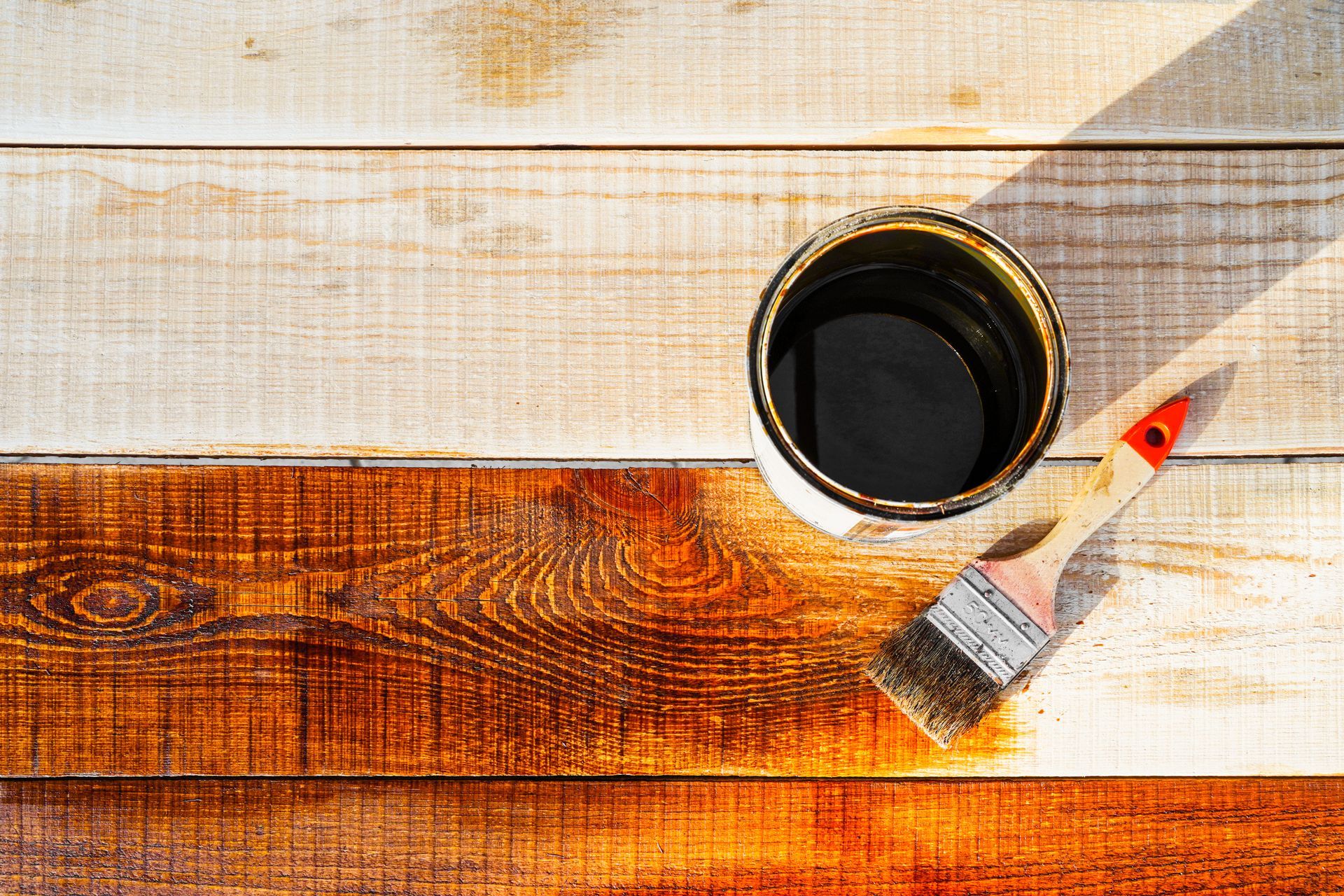 A paintbrush resting beside an open can of dark wood stain on half-finished, light-colored wooden planks.