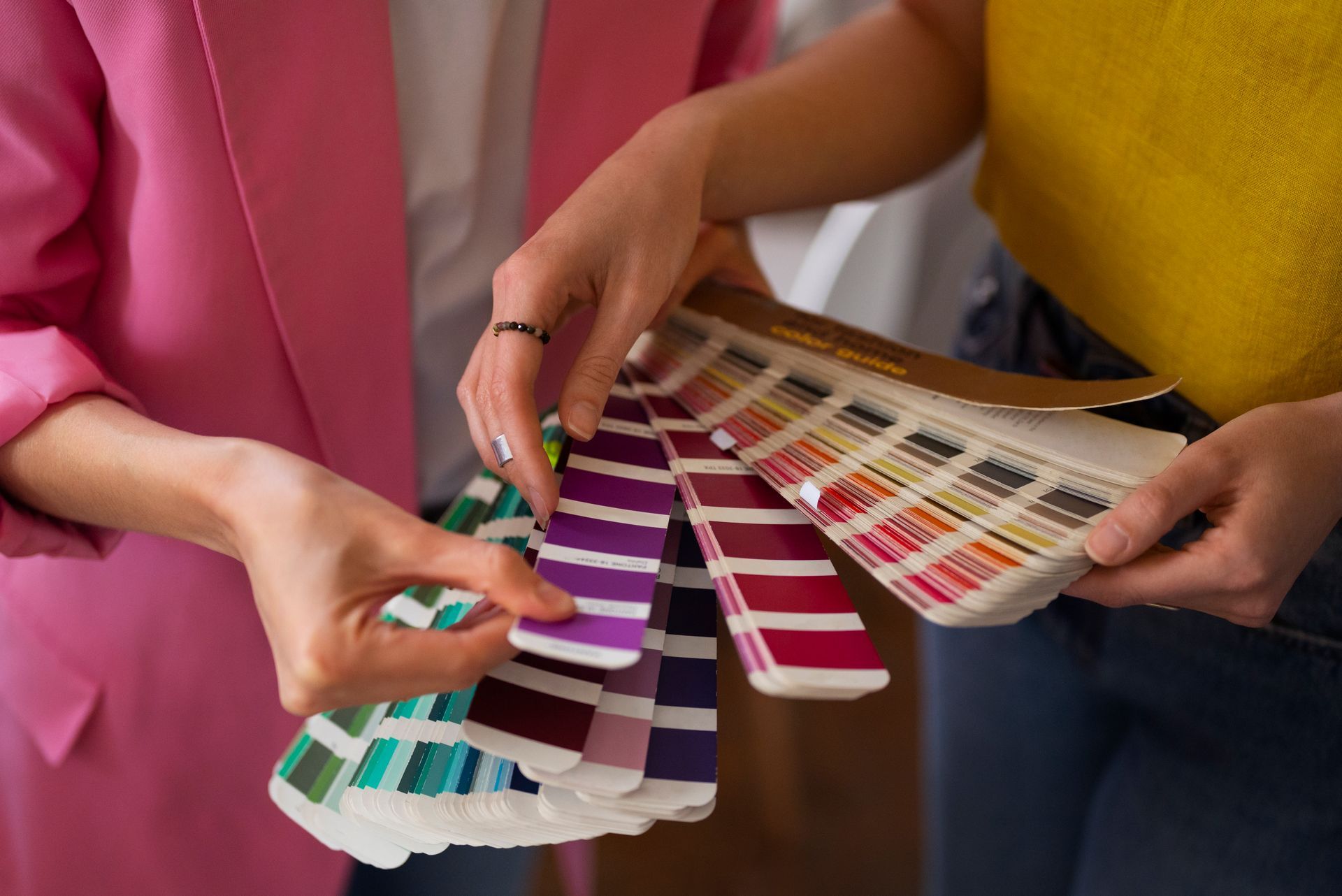 Two people examining color swatches, indoors. One in pink blazer, the other in yellow top.