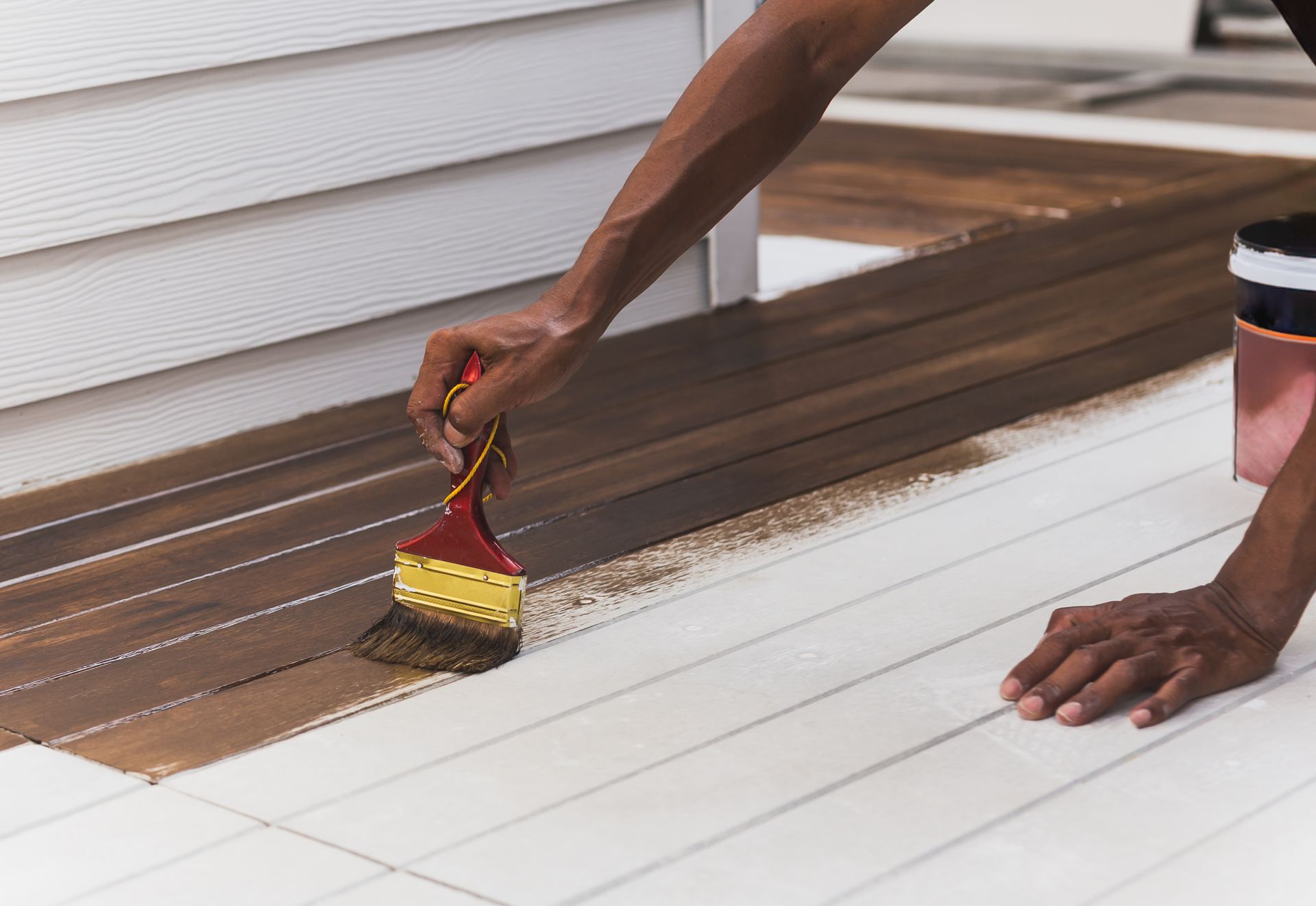 Person staining a wooden deck with a brush. The deck is partly stained brown; the other portion is white.