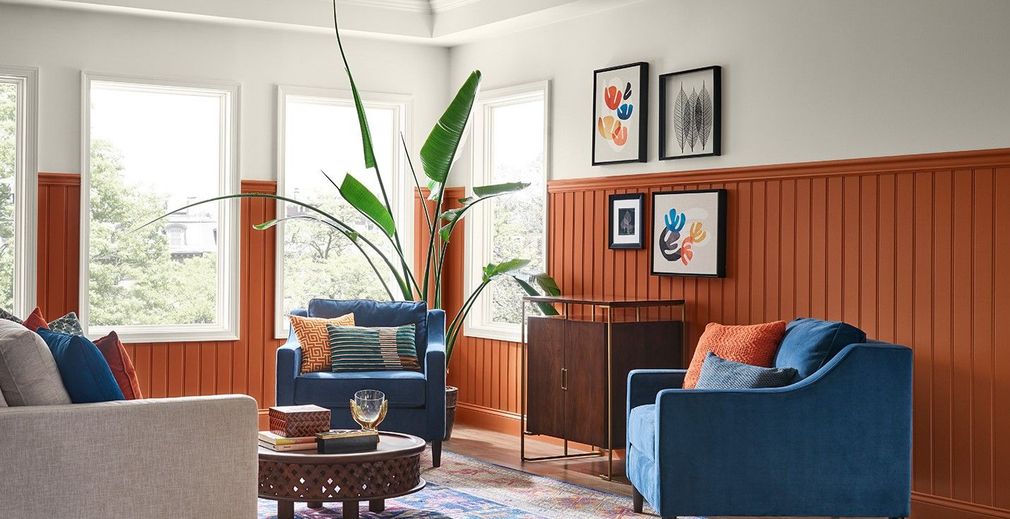Cozy living room with blue chairs, wood wainscoting, large windows, and a tall potted plant