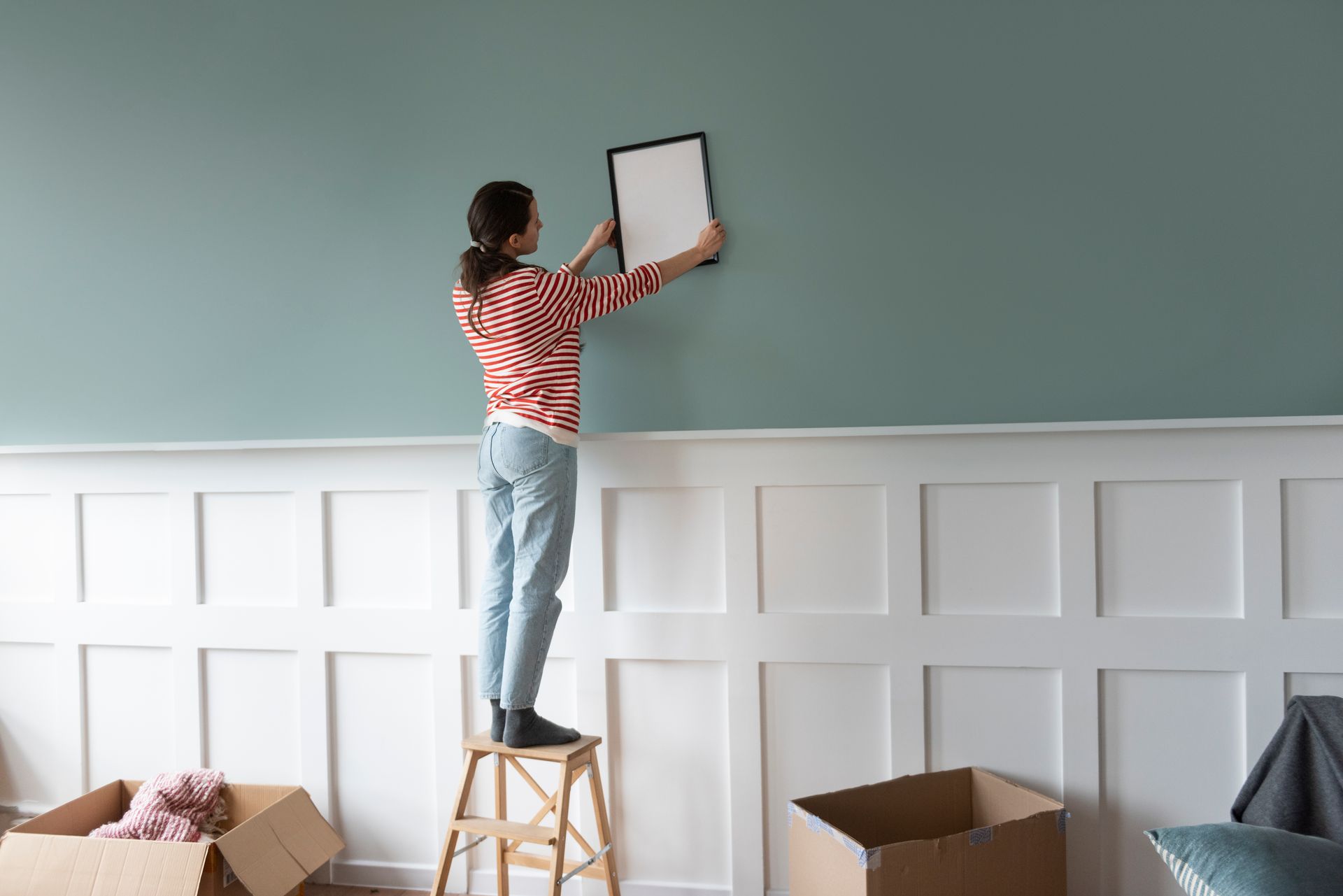 Woman on step stool hanging a picture on a teal wall above white paneling. Cardboard boxes sit below.
