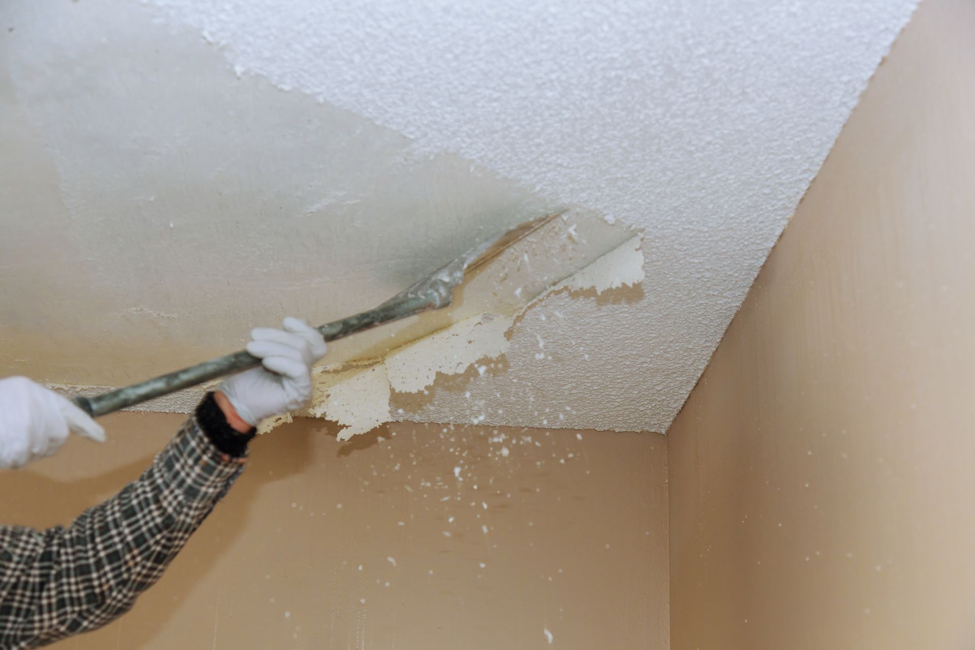 Person scraping textured ceiling with a scraper tool. Drywall dust falls, beige walls in corner.