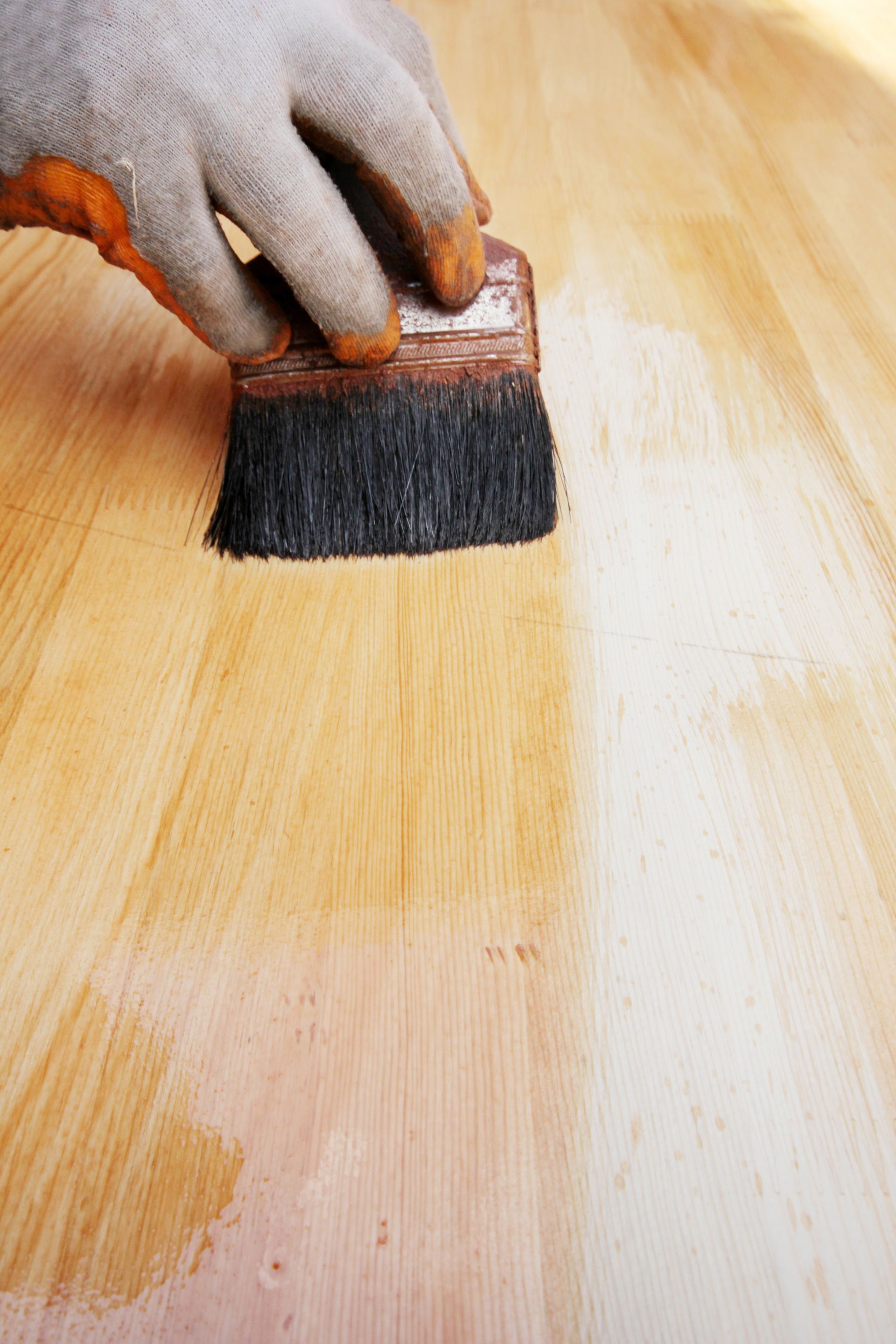 Gloved hand brushing stain onto a wooden board, leaving a light amber finish