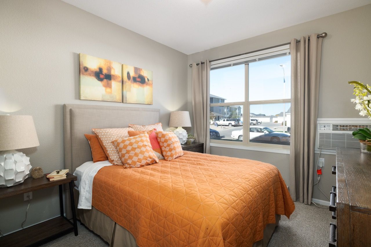 Furnished bedroom in an apartment featuring an orange quilt, pillows, two nightstands with lamps, and a large window.