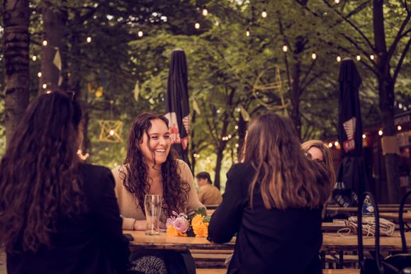 Three people sit at an outdoor wooden table in a tree-lined area decorated with hanging string lights.