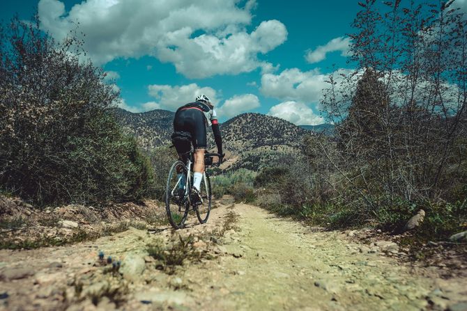 A cyclist riding a bike up a dirt path through a hilly, arid landscape under a blue sky with scattered clouds.
