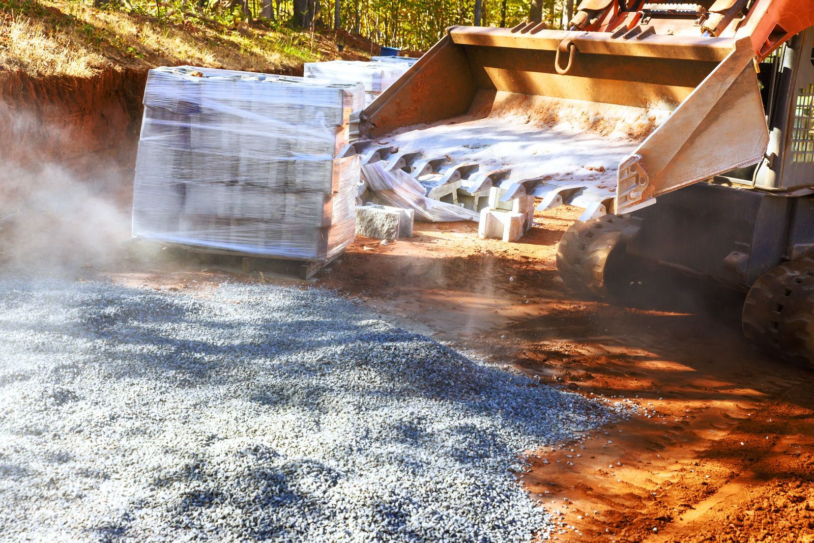 Skid steer spreading gravel on a dirt road. Pallets of wrapped bricks are in the background.