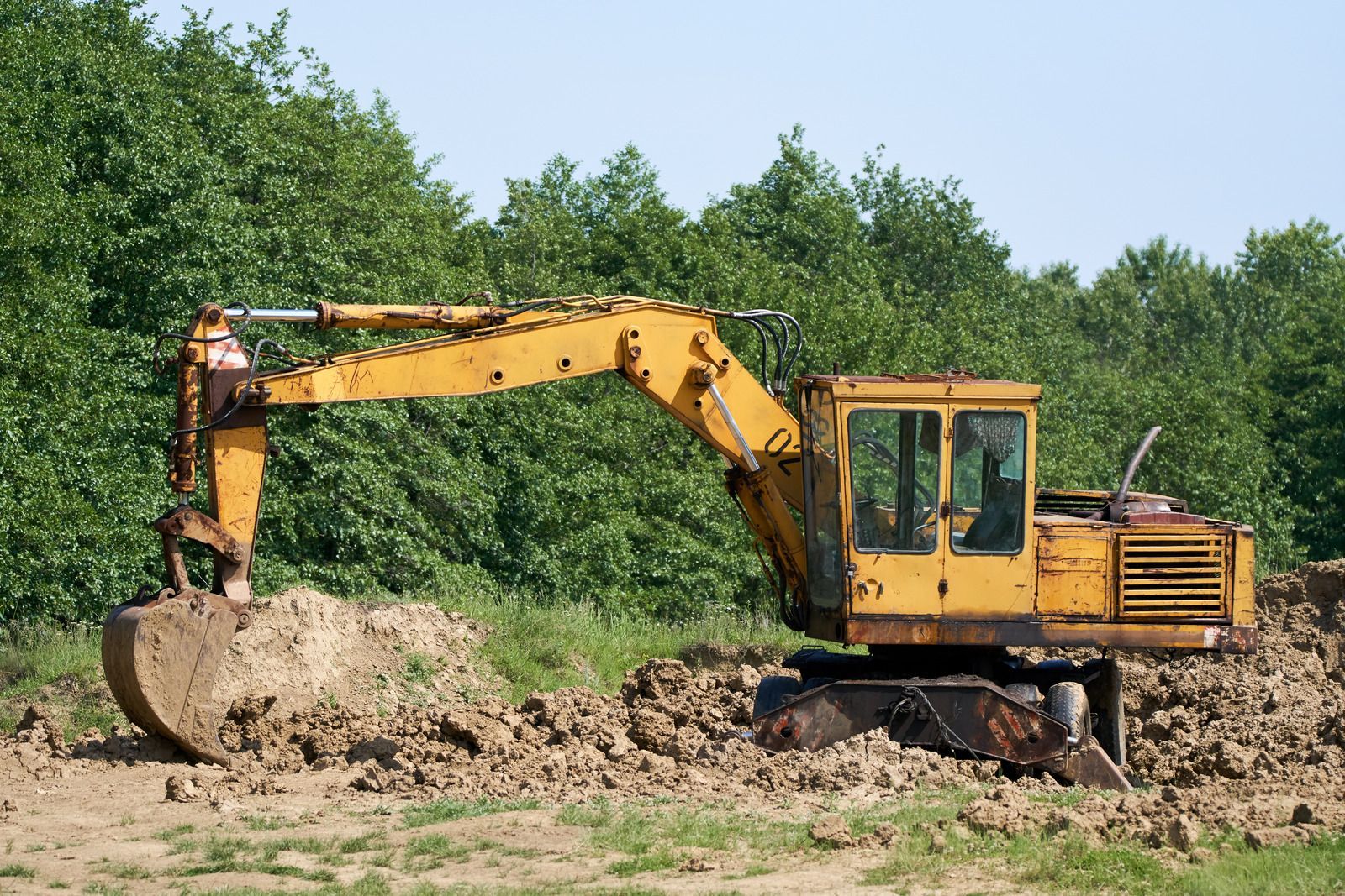 Yellow excavator digging in a dirt pile, with green trees in the background.