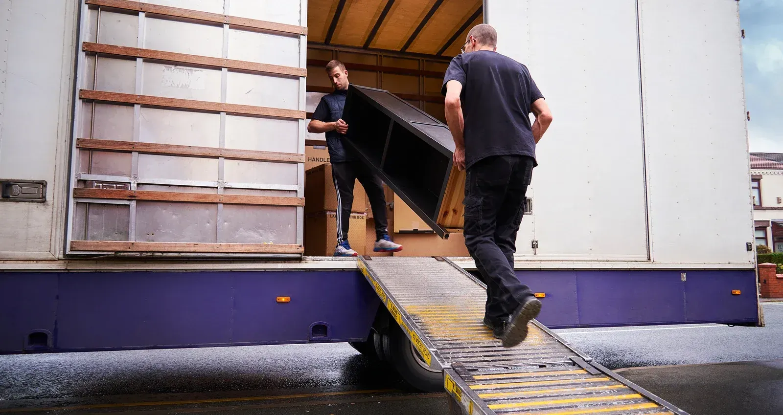 Two movers unloading a cabinet from a truck onto a ramp, outdoors.