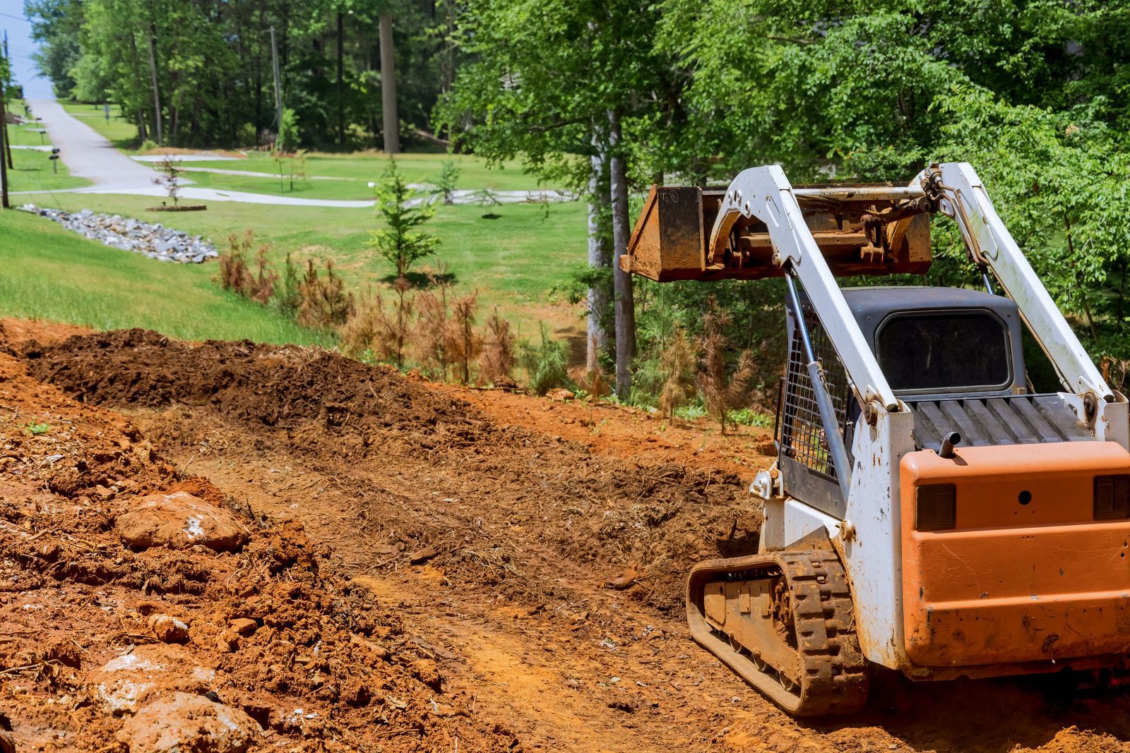 Orange skid steer loader excavating dirt on a slope, with a distant road and greenery.