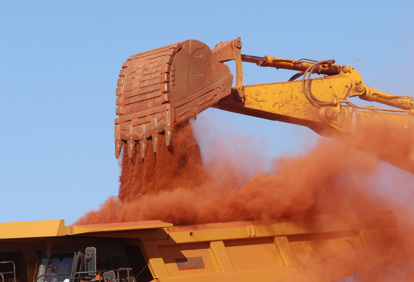 Yellow mining dump truck loaded with ore, being filled by an excavator. Desert setting, bright blue sky.