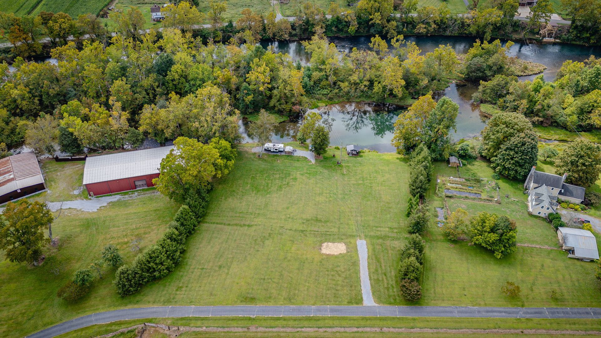 Aerial view of a grassy field next to a river and a road. Barn, trees, and a house are visible.