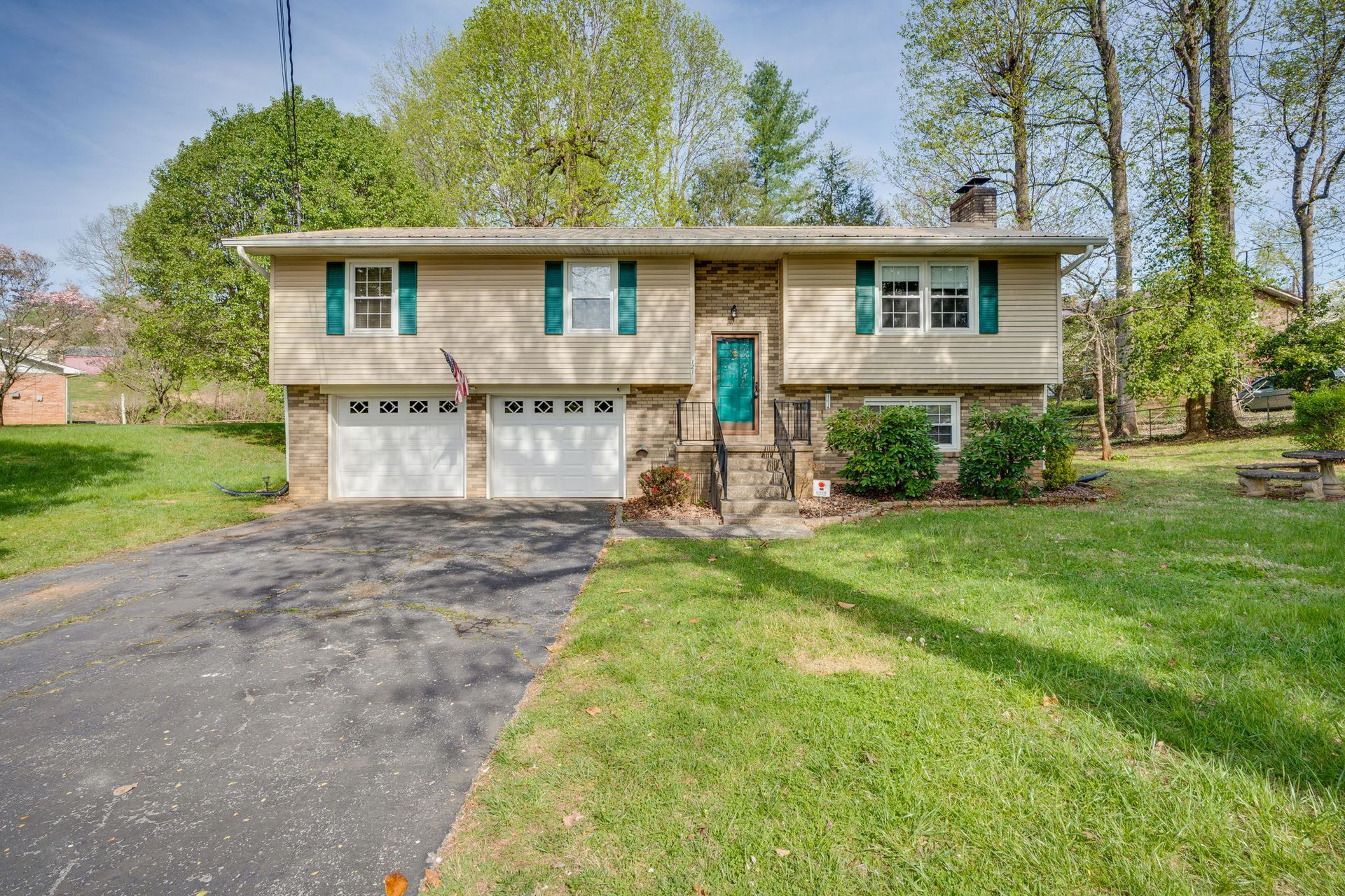 A beige split-level house with a two-car garage, green front door, and a paved driveway, set on a grassy, tree-lined lot.