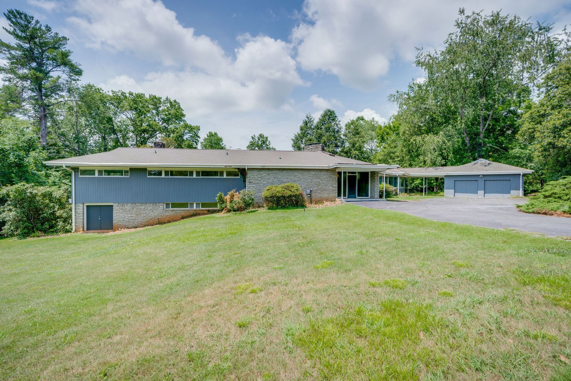 Single-story ranch house with large front lawn, trees, and a detached garage on a cloudy day