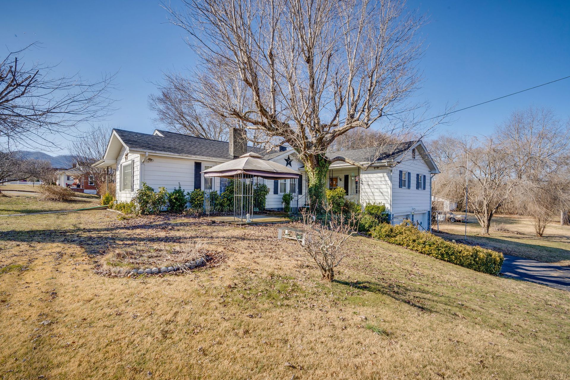 White house with a gazebo, bare tree in front, on a sunny, dry lawn.