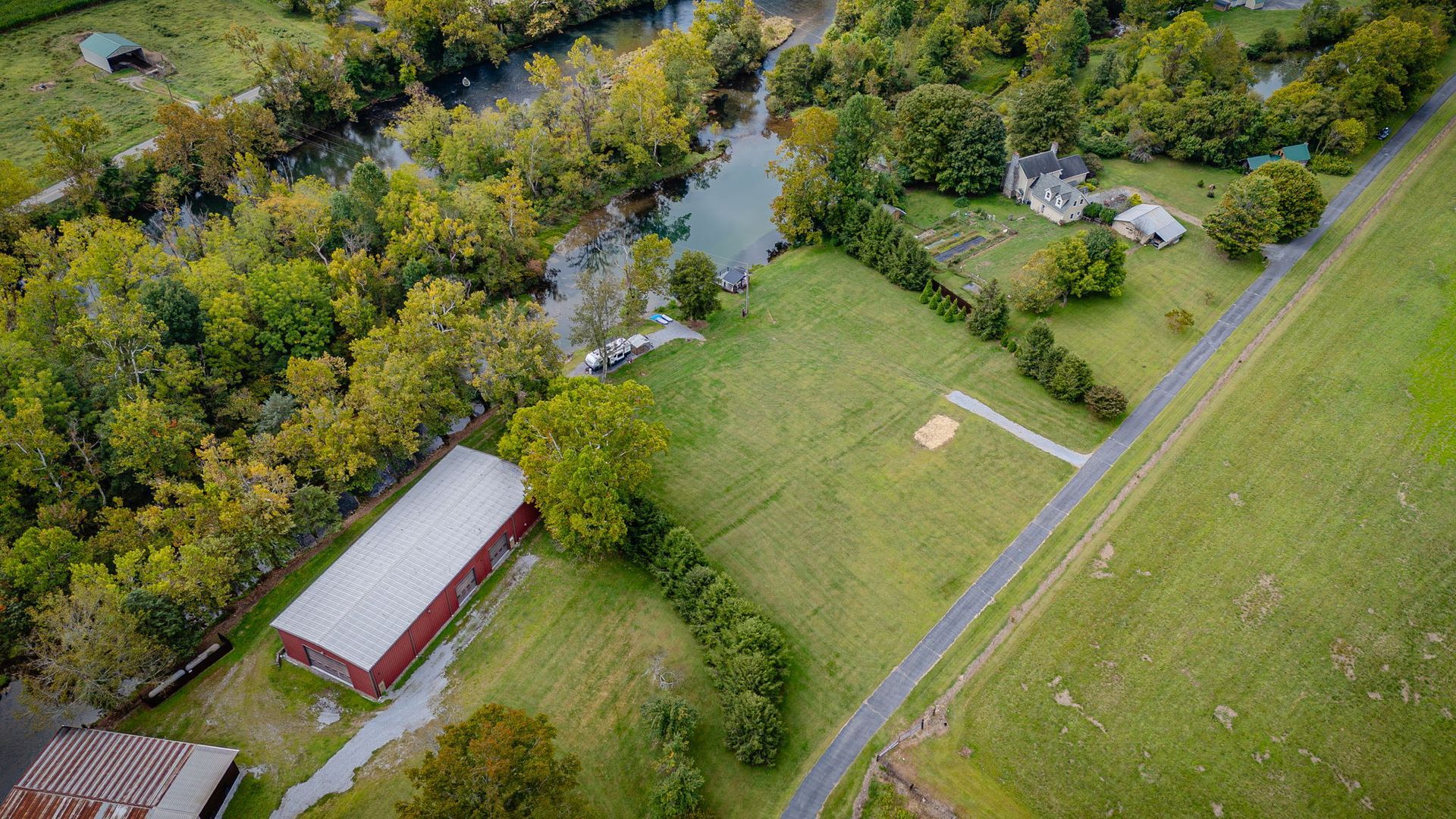 Aerial view of a red barn, houses, trees, and a road alongside a creek in a green field.