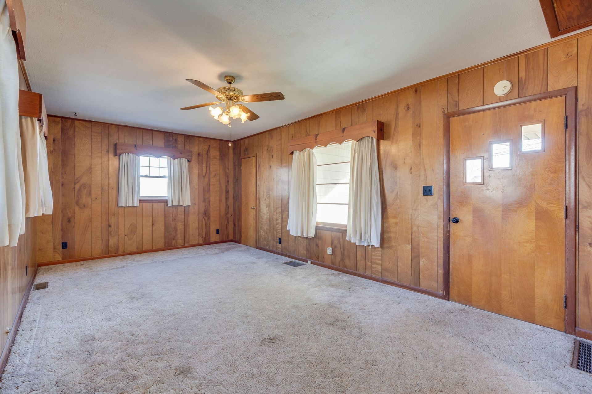 Empty room with wood paneling, beige carpet, two windows, and a ceiling fan.