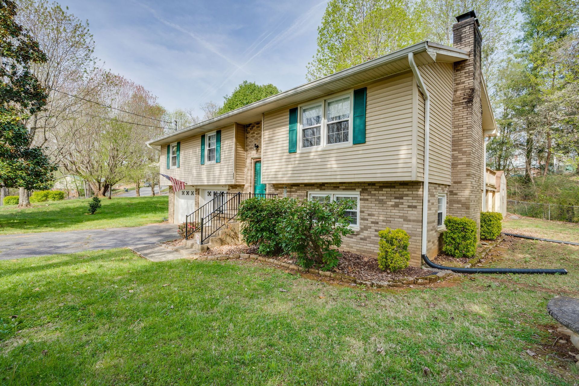 A two-story house with tan siding and stone masonry, situated on a grassy, tree-lined lot under a bright blue sky.
