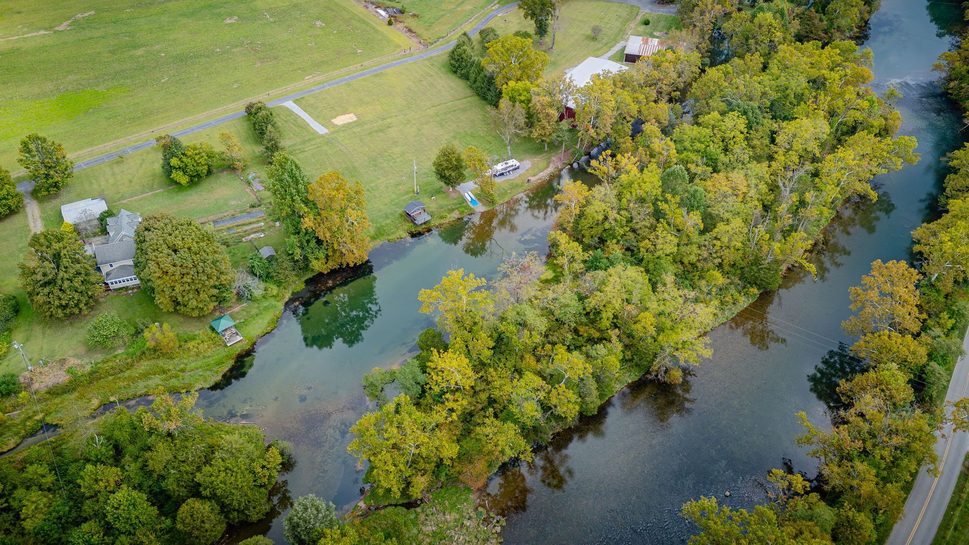 Aerial view of a river winding through lush green trees, near a grassy field and buildings.