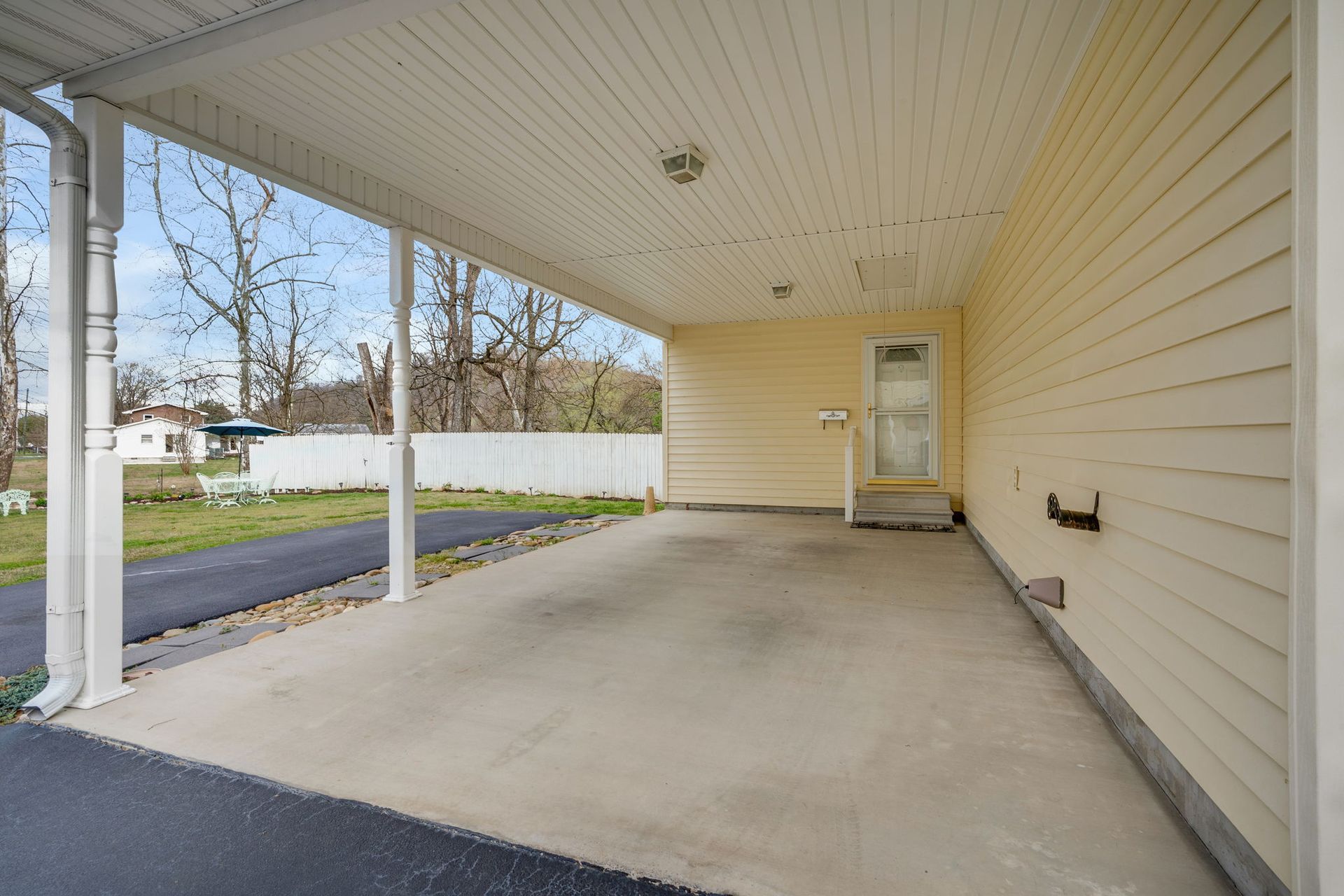 A beige home exterior with a covered carport featuring a concrete floor, white support posts, and a door to the house.