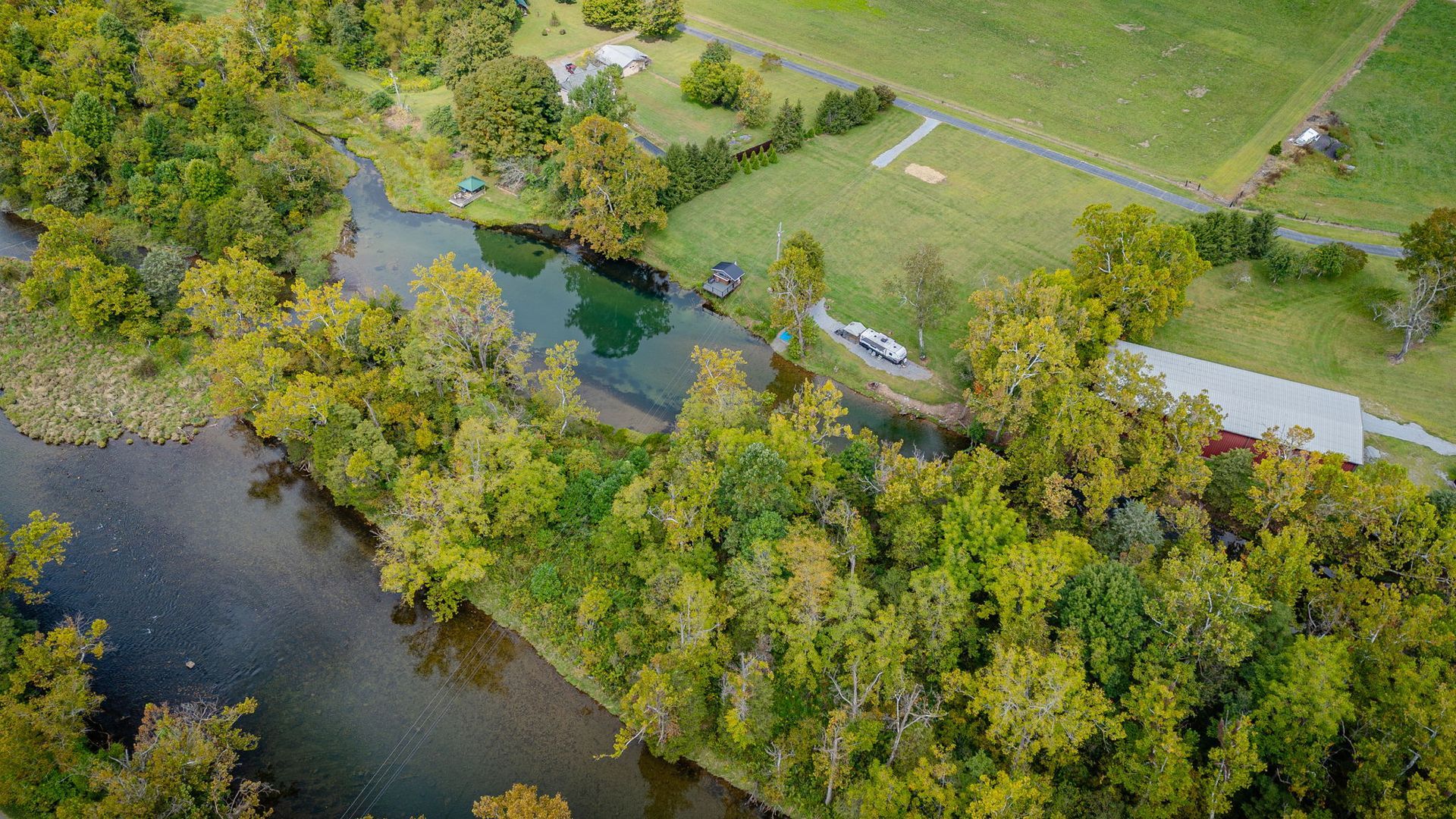 Aerial view of a river winding through lush green trees with a grassy field and buildings on the right.