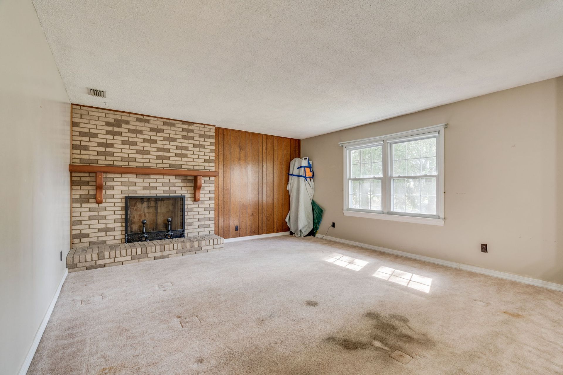 A living room featuring a stone fireplace, wood paneling, a large window, and light carpet with a dark stain.