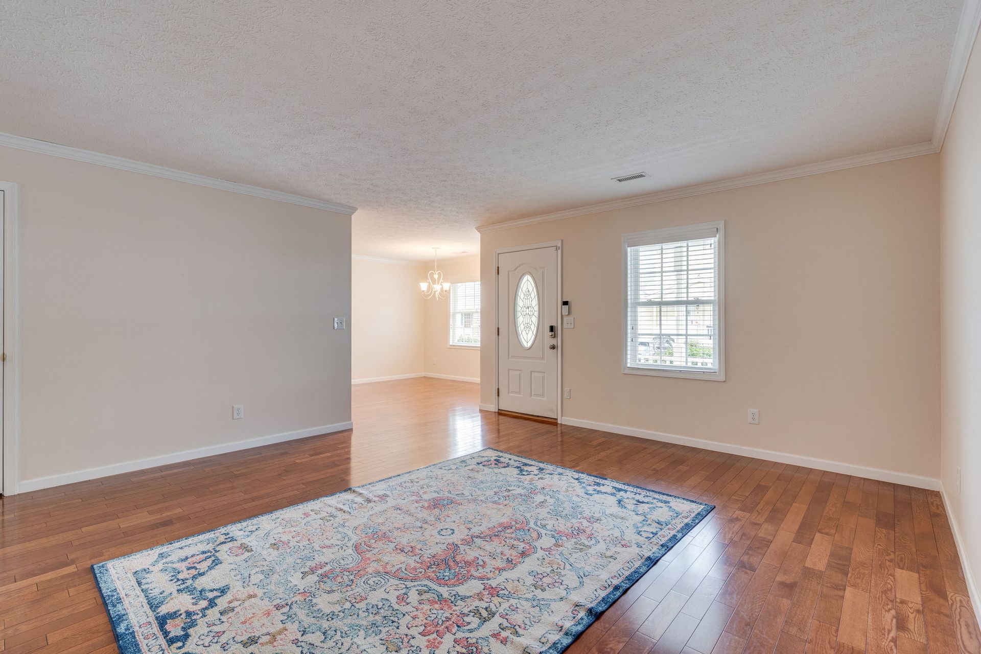 An empty living room with hardwood floors, a patterned area rug, a white front door, and a glimpse of a dining area.