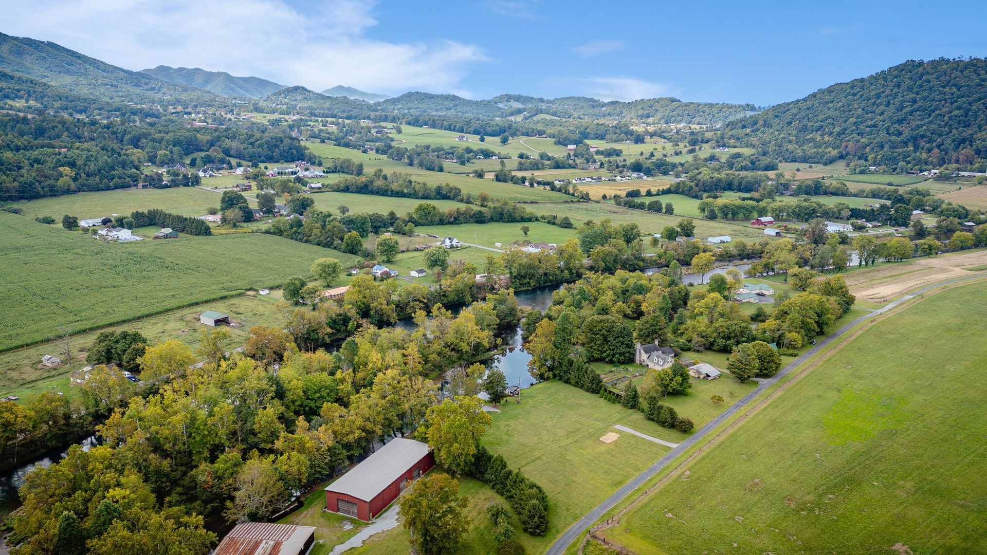 Scenic aerial view of a valley with a river, green fields, trees, and houses, under a blue sky.