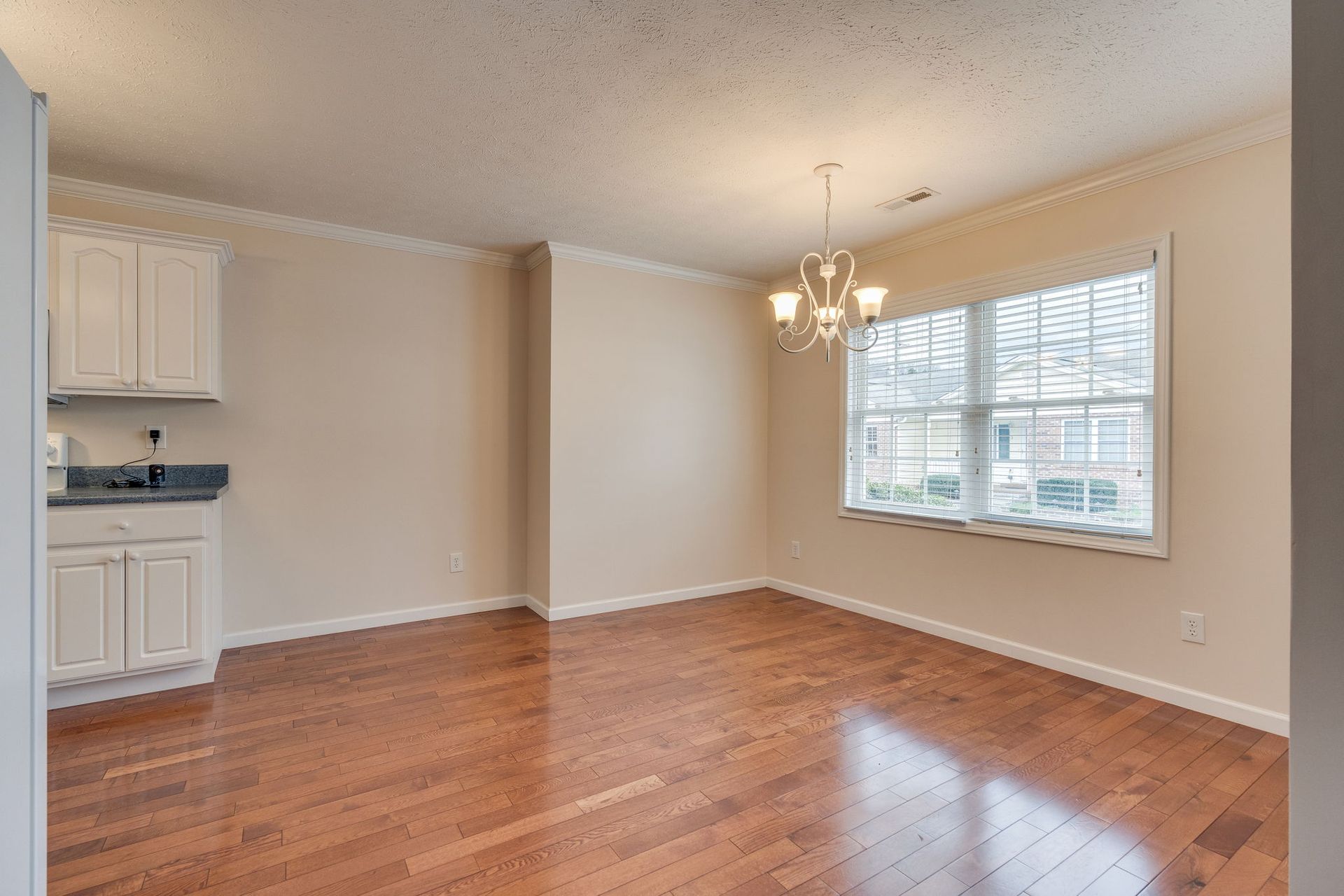 A brightly lit dining area featuring hardwood floors, off-white walls, a white kitchen cabinet, and a glass chandelier.