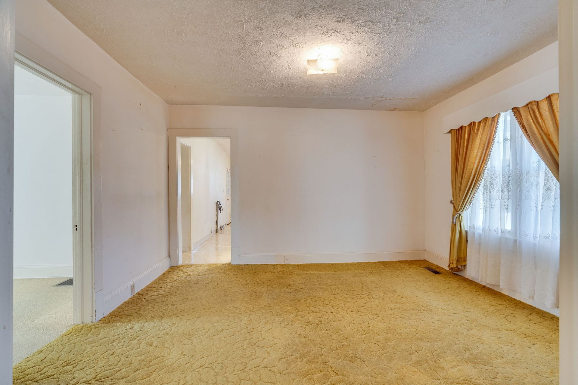 Empty room with gold carpet, white walls, and a window with curtains; a doorway visible on left.