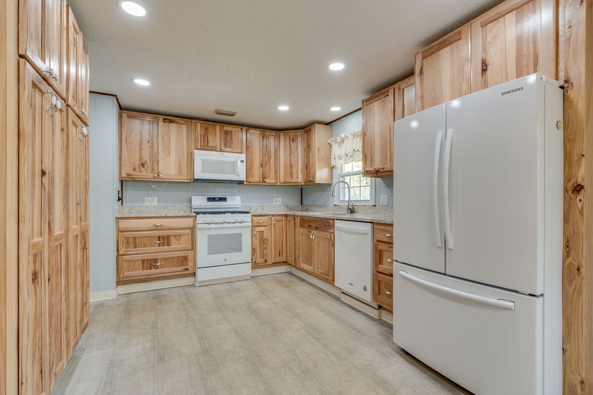 A rustic kitchen with light wood cabinets, white appliances, and wood-patterned flooring.