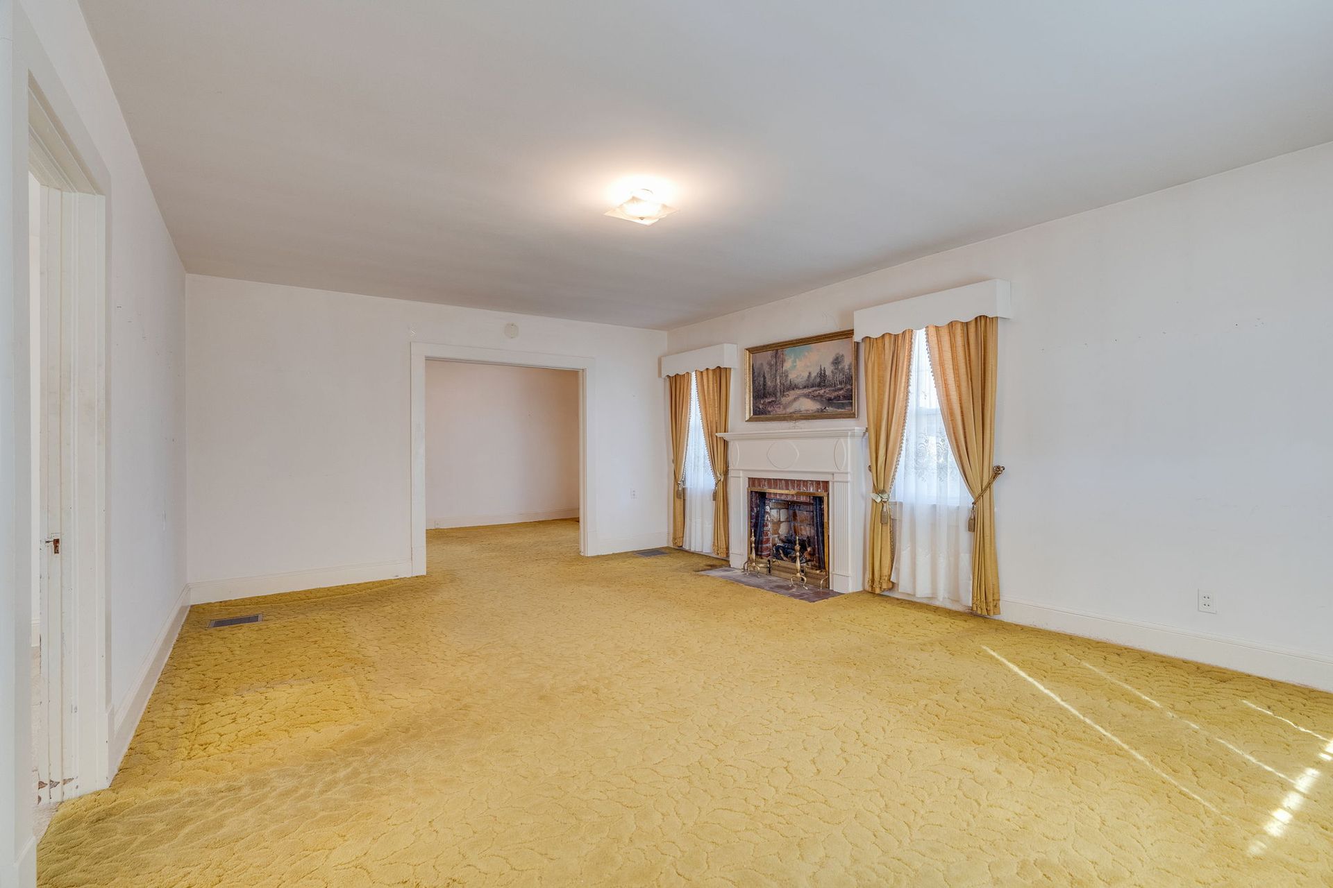 Empty living room with yellow carpet, white walls, fireplace, and window with curtains.