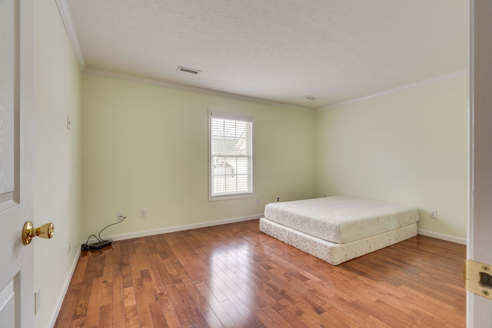 A sparse, empty bedroom with light green walls, hardwood floors, and a single mattress on the floor by a window.