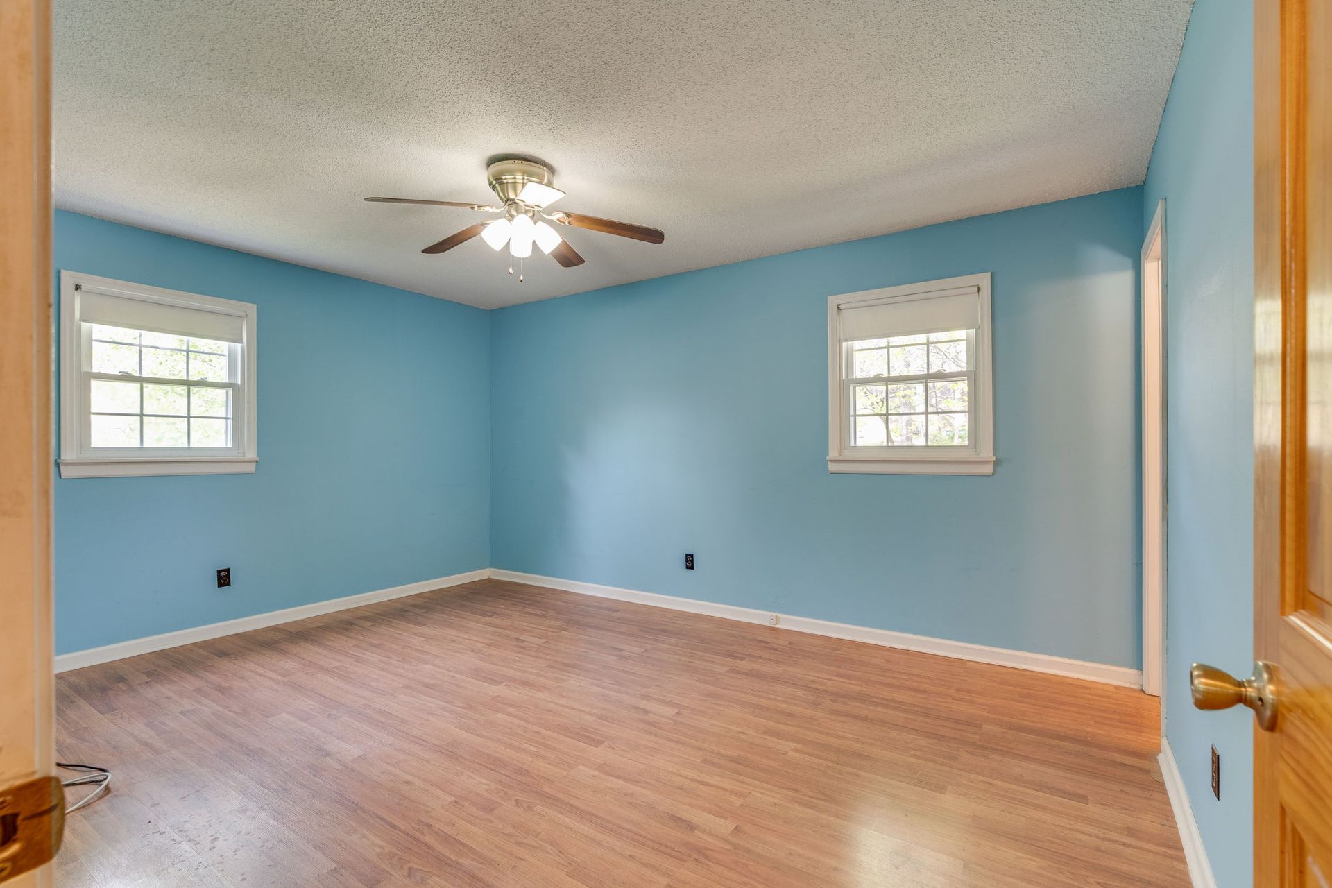 An empty bedroom with light blue walls, wood-look flooring, two windows with white blinds, and a ceiling fan.