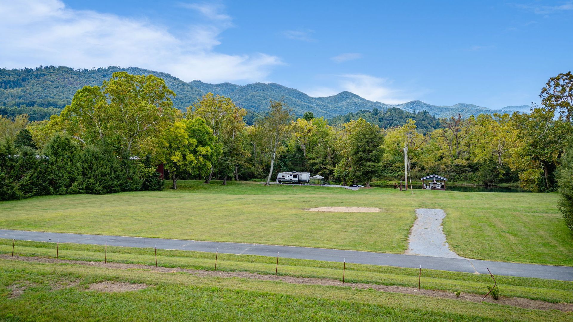 Grassy field with a gravel path, trees, and mountain backdrop on a sunny day.