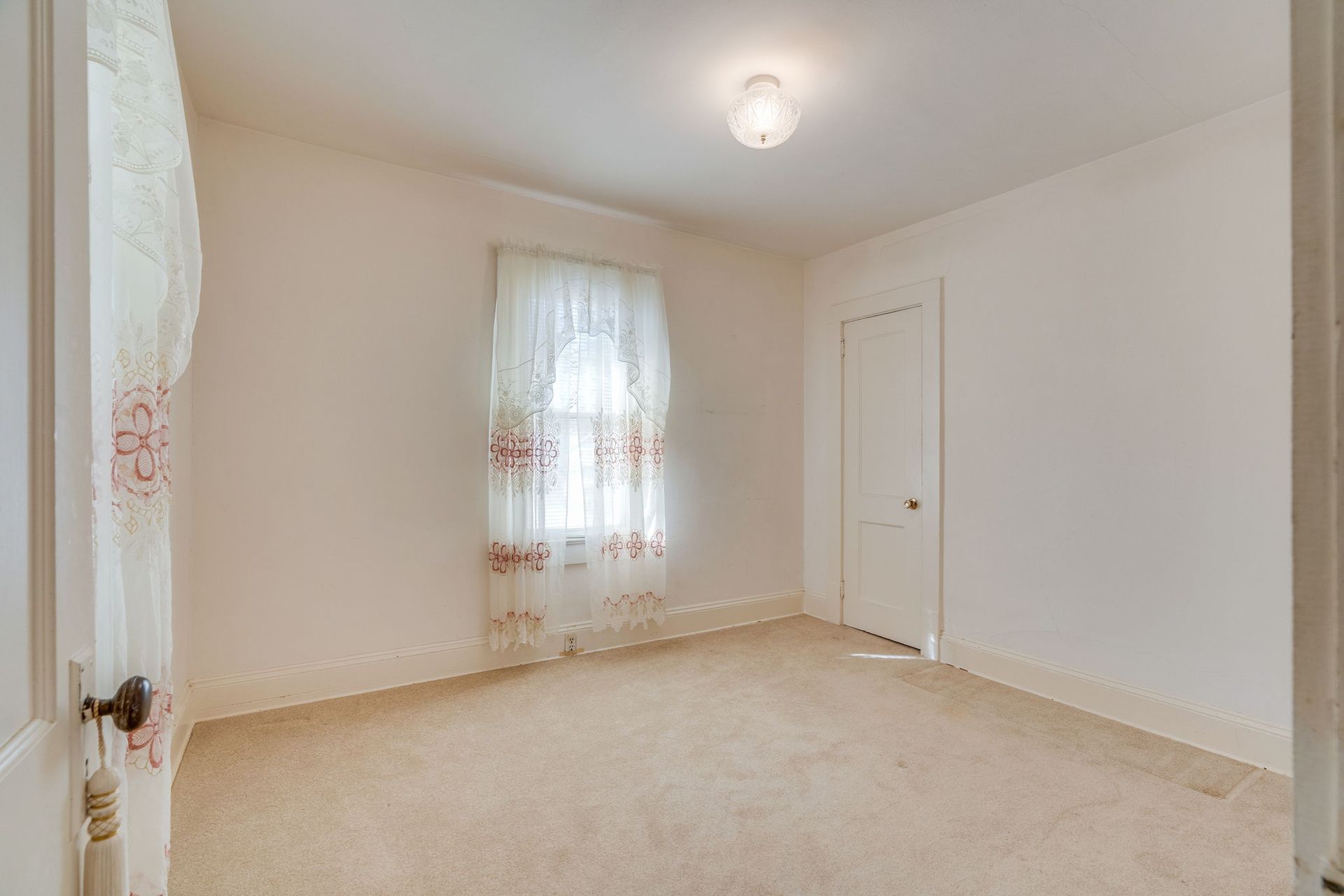 Empty bedroom with beige carpet, a small window with lace curtains, and a white door.