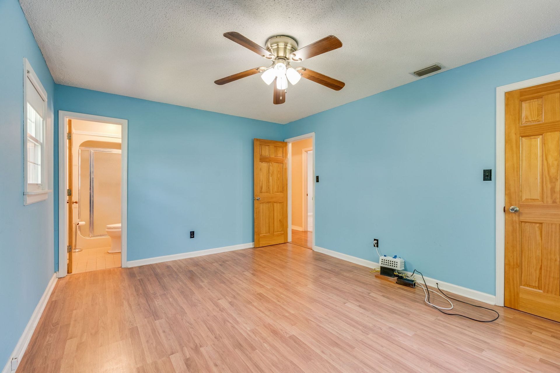 An empty bedroom with light blue walls, wood-look flooring, a ceiling fan, and two wooden doors.
