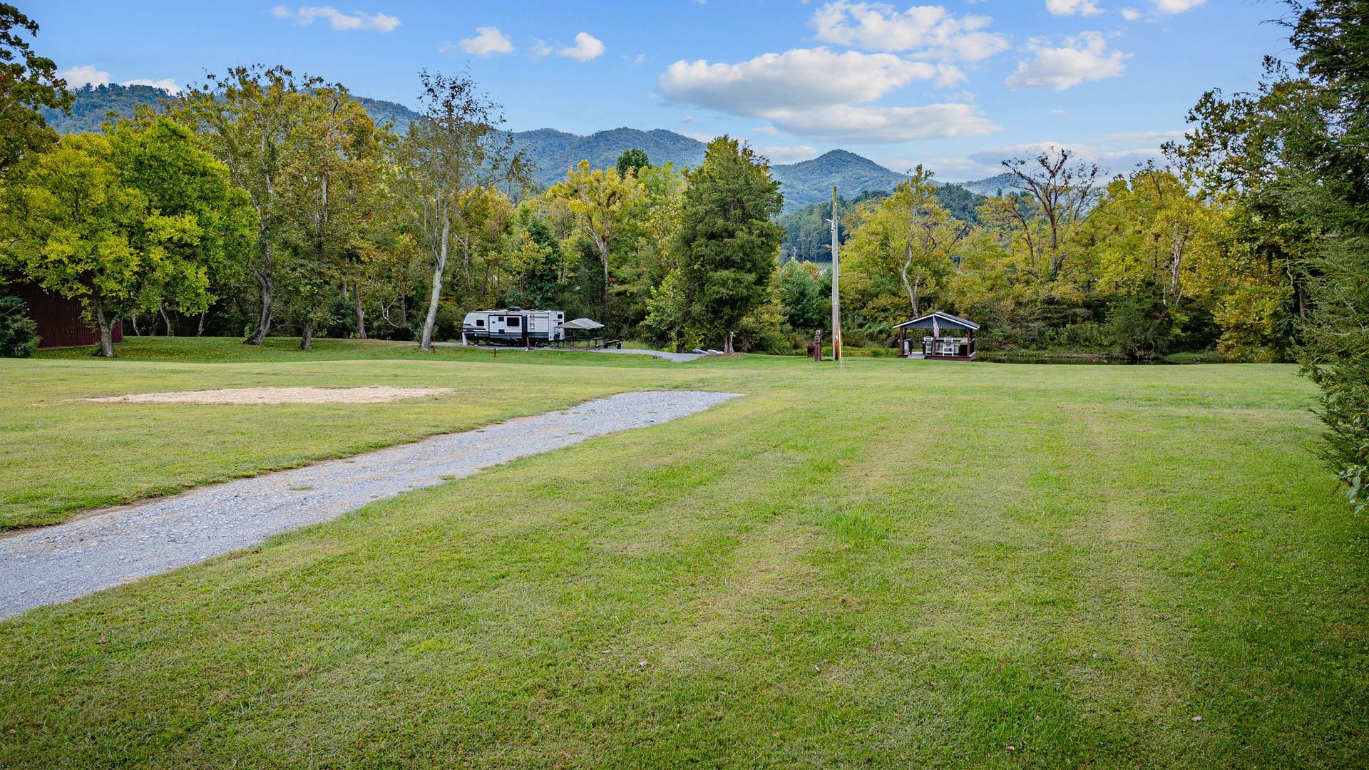 A grassy field with a gravel path, trees, and mountains in the distance under a blue sky.