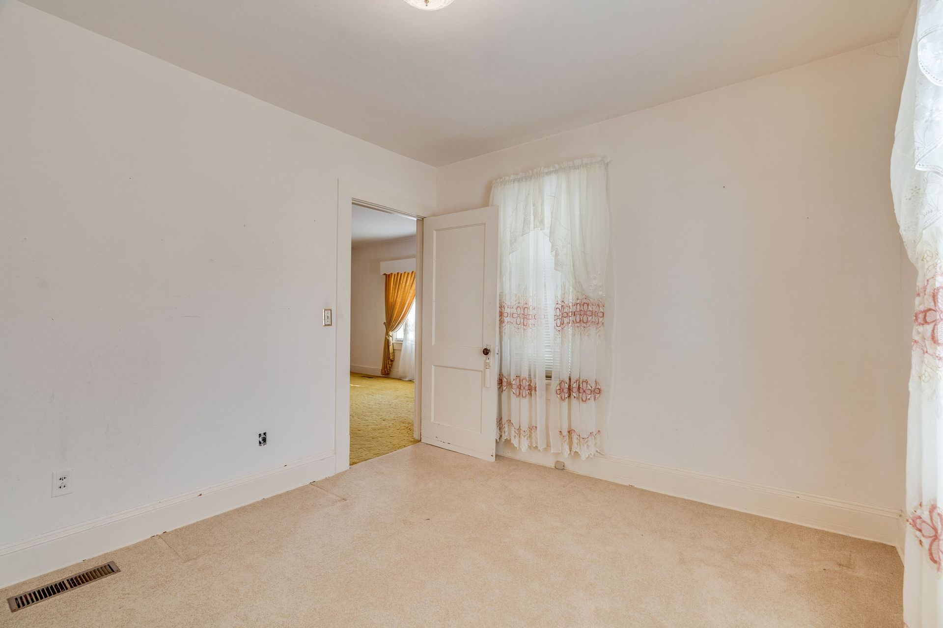 Empty room with cream-colored walls, carpet, and an open doorway to another room. A window with a curtain is on the right.
