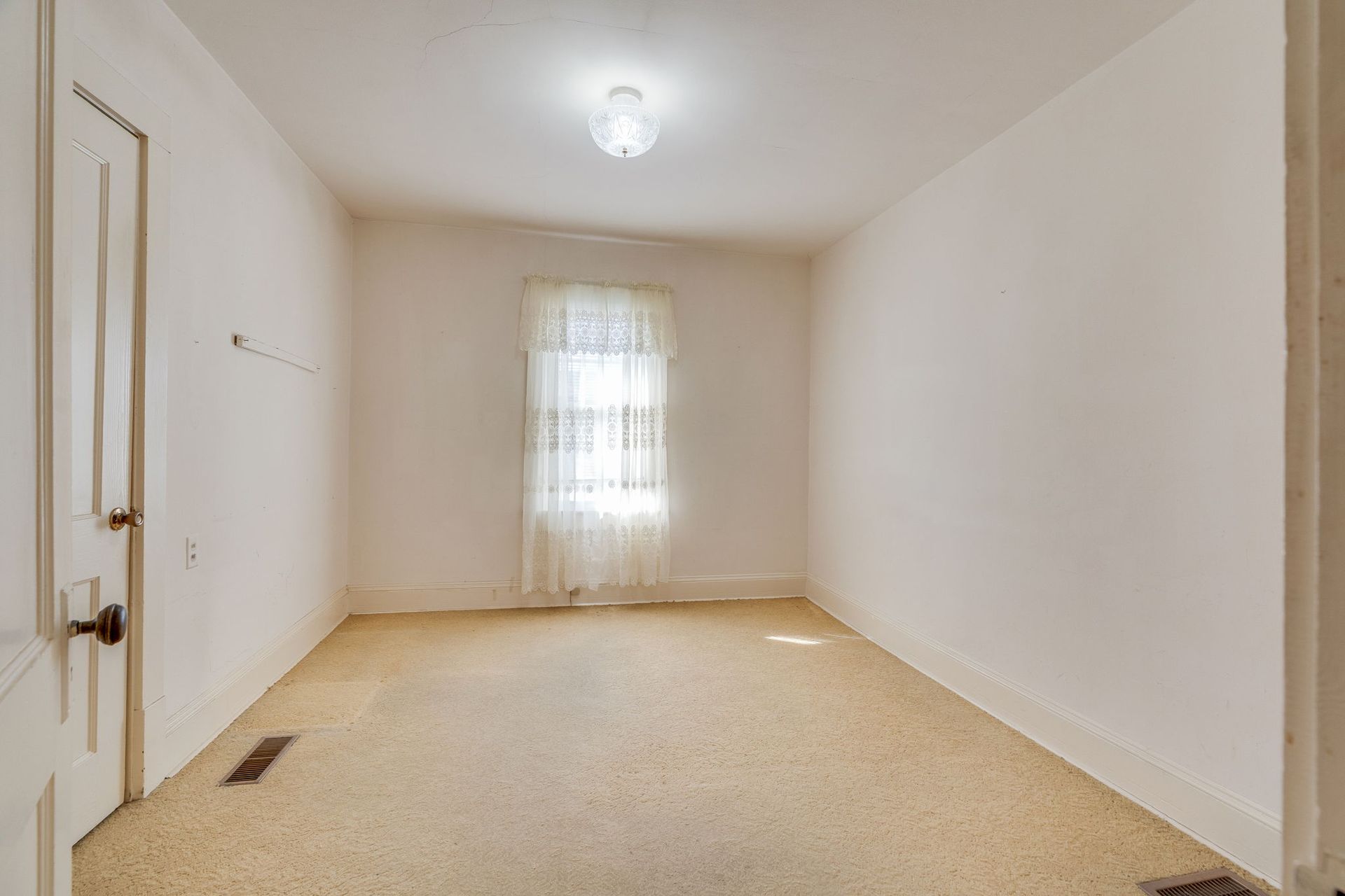 Empty room with off-white walls, carpet, and a window with a lace curtain. Door is on the left.
