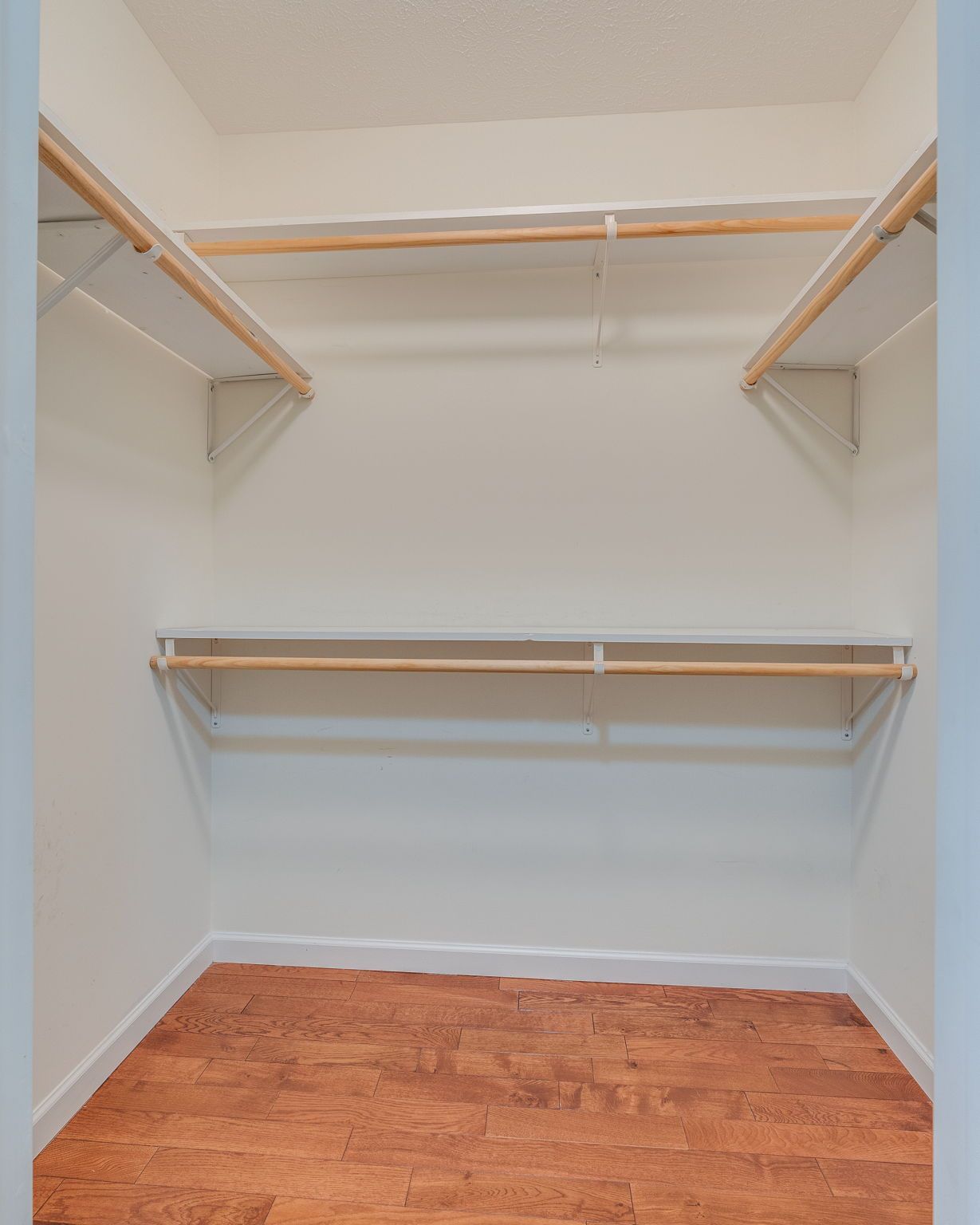 An empty walk-in closet with white walls, wood-look flooring, and two levels of white shelving with wooden clothes rods.