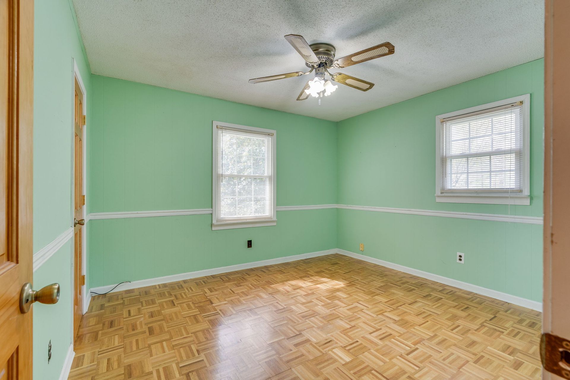 An empty room with mint green walls, chair rail molding, light wood parquet flooring, two windows, and a ceiling fan.