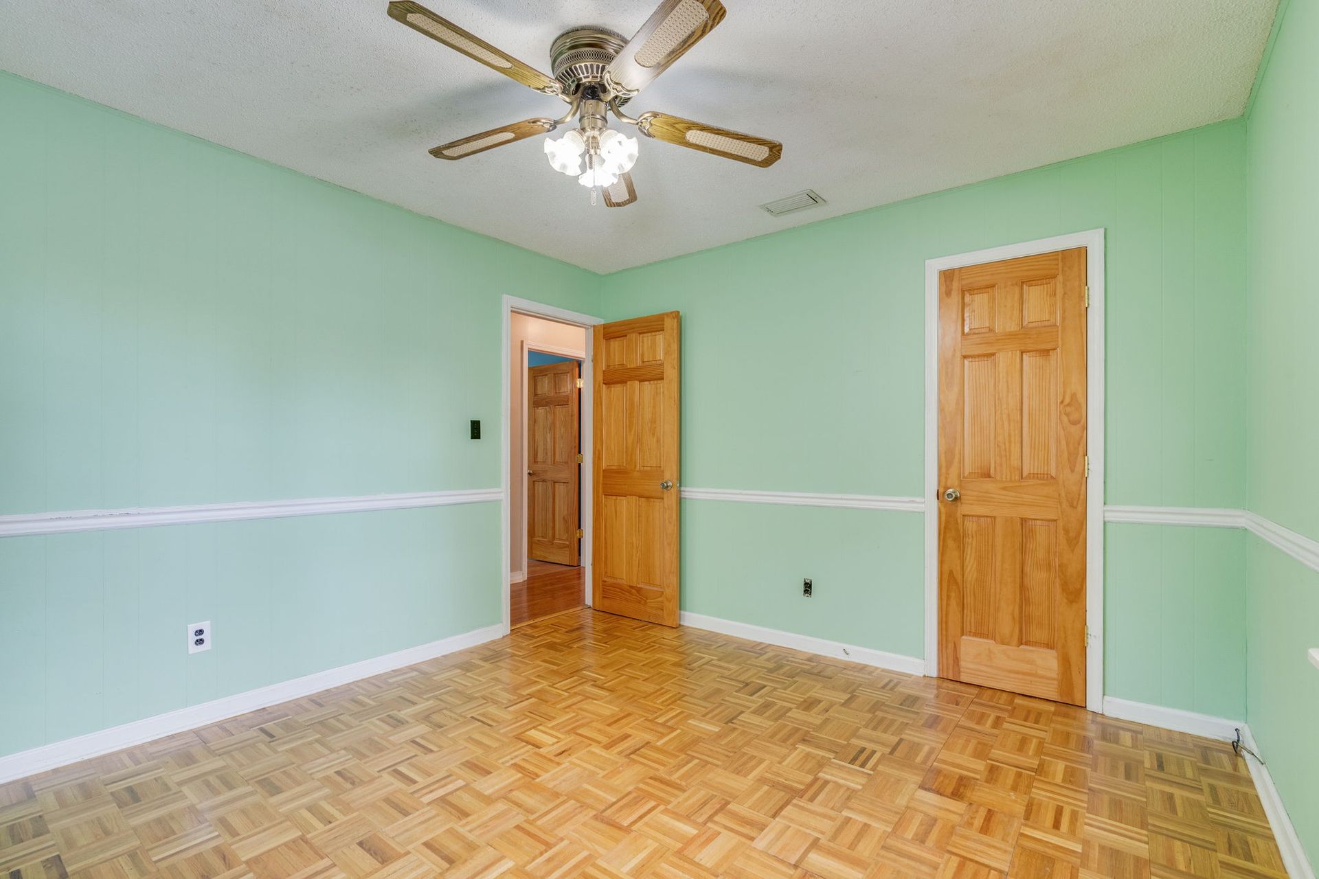 A bright, empty room with light green walls, a white chair rail, parquet wood flooring, and a ceiling fan.