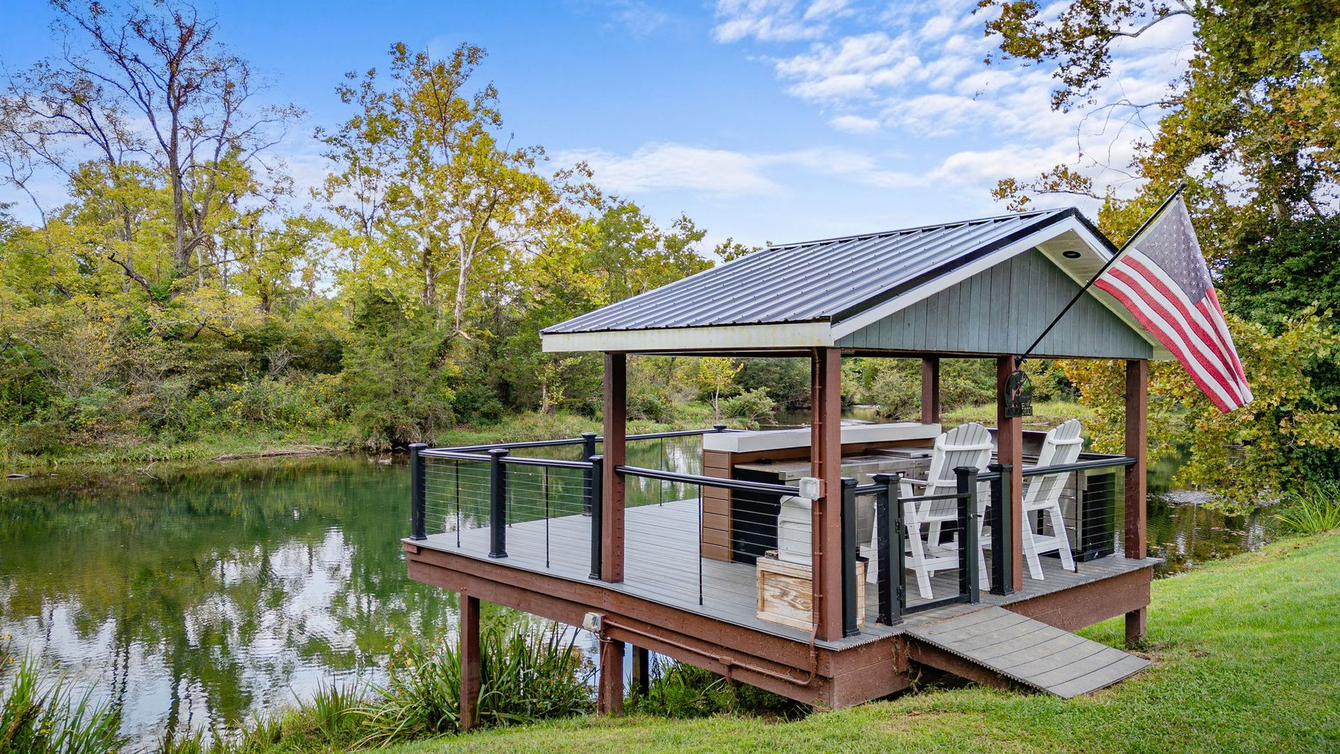 Gazebo deck over water with American flag, white chairs, and trees in background.