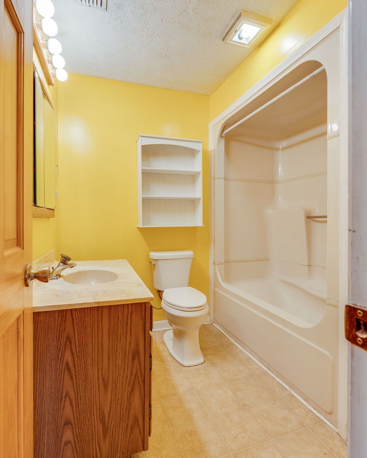 A small bathroom with yellow walls, a wooden vanity, white toilet, wall-mounted white shelves, and a shower-tub unit.