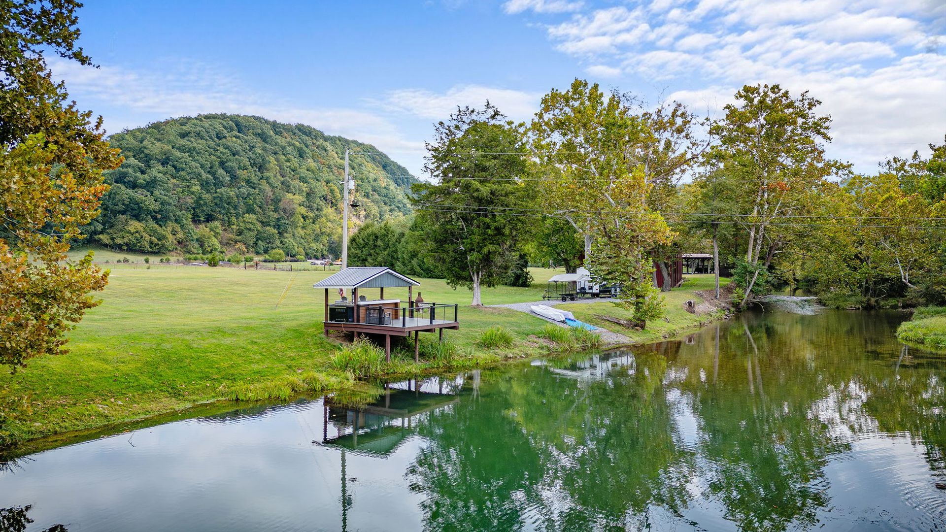 A wooden dock on a calm river, green field, trees, and a distant mountain under a blue sky.
