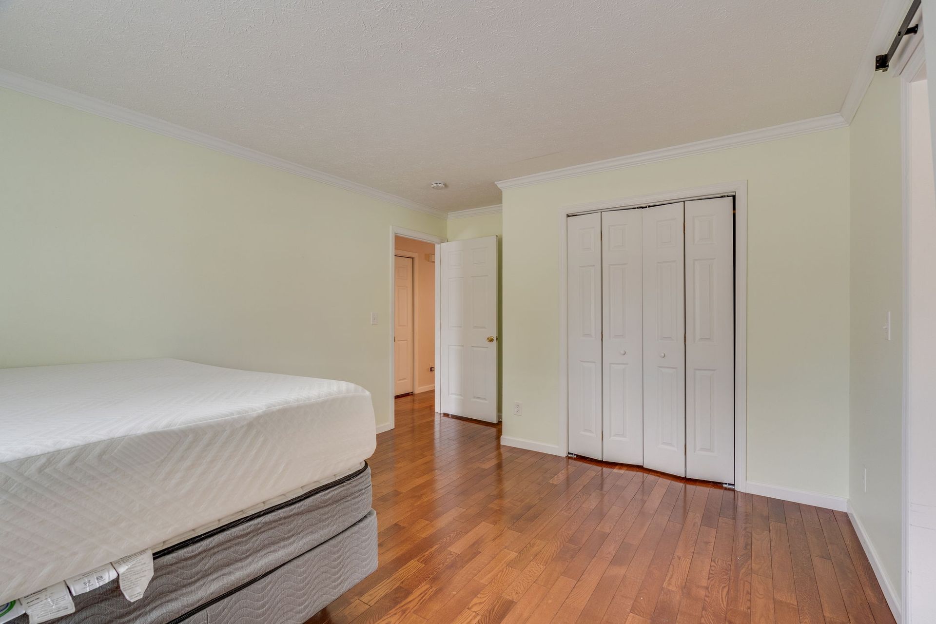 A bedroom with light walls and hardwood floors, featuring a bed mattress and white bi-fold closet doors.