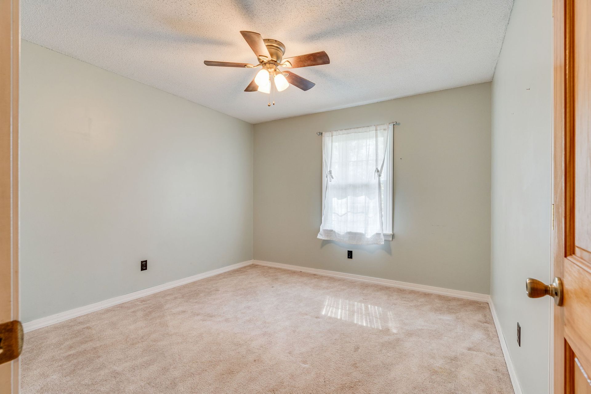 An empty bedroom with light gray walls, tan carpeting, a ceiling fan, and a window with white curtains.