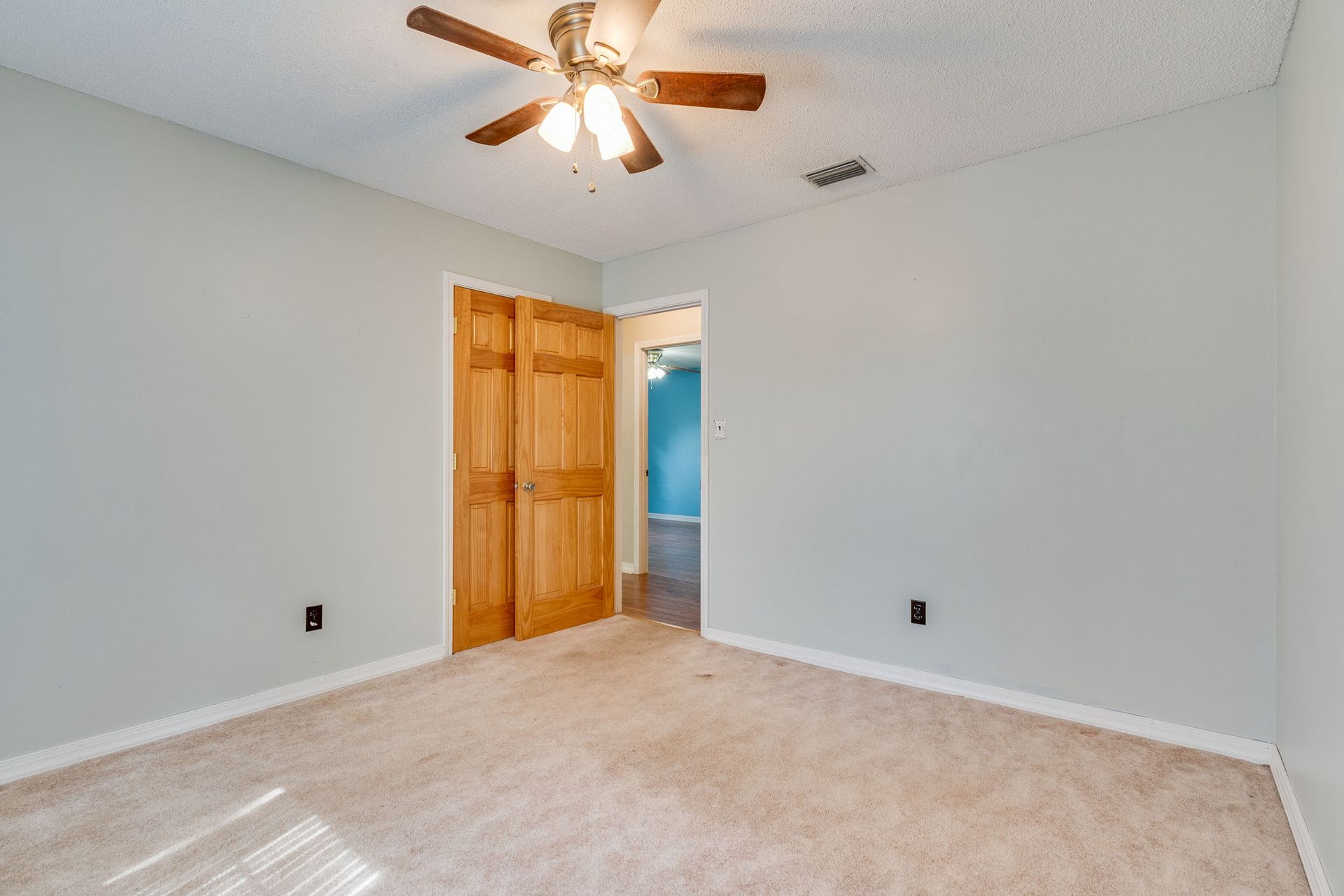 An empty bedroom with light gray walls, tan carpet, a ceiling fan, and wooden double doors leading to a hallway.